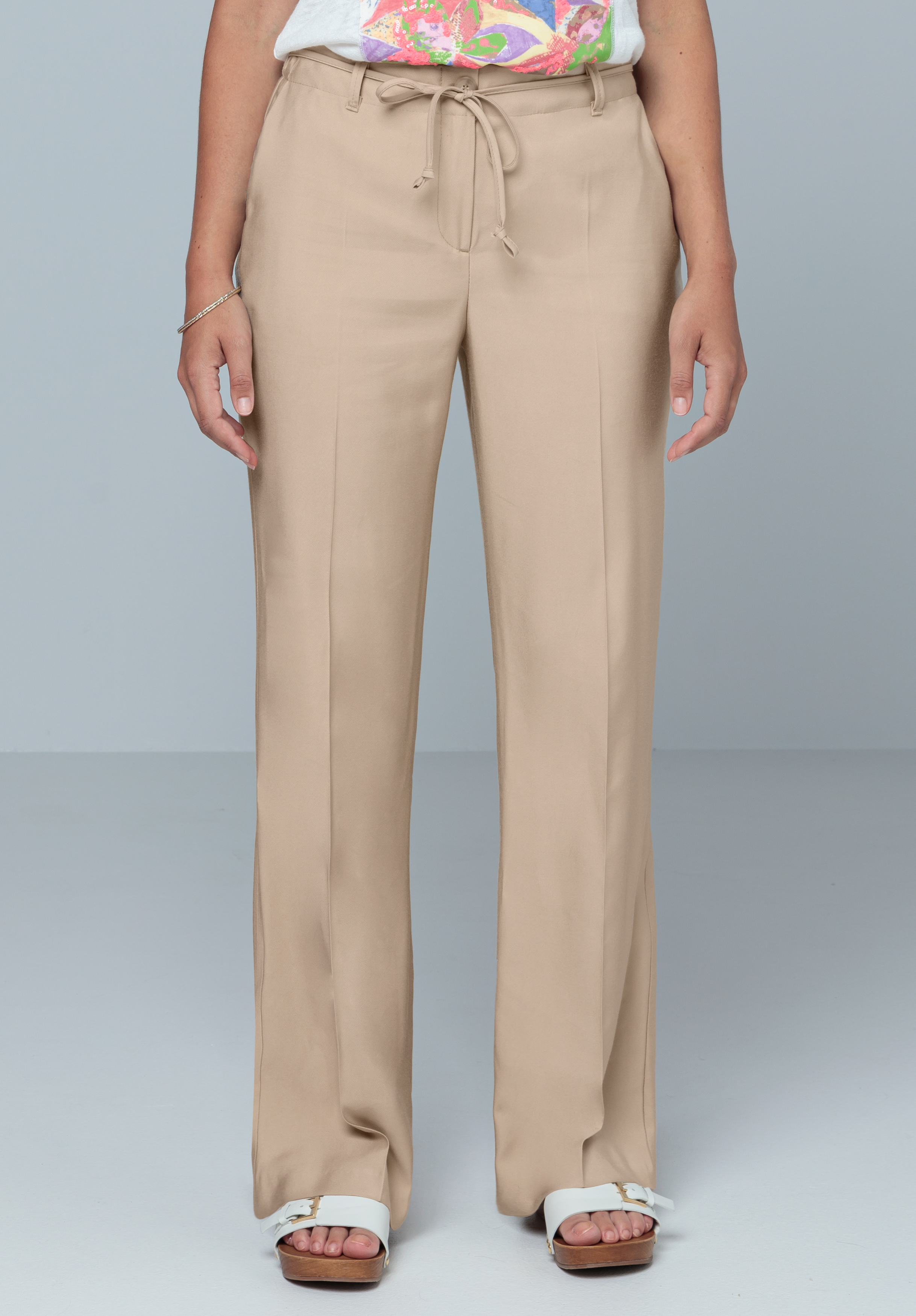 Eine Person, die beige Hosen mit weitem Bein und Kordelzug in der Taille, ein weißes T-Shirt, weiße Sandalen mit offenen Zehen und ein Armband trägt, steht vor einem schlichten hellgrauen Hintergrund.