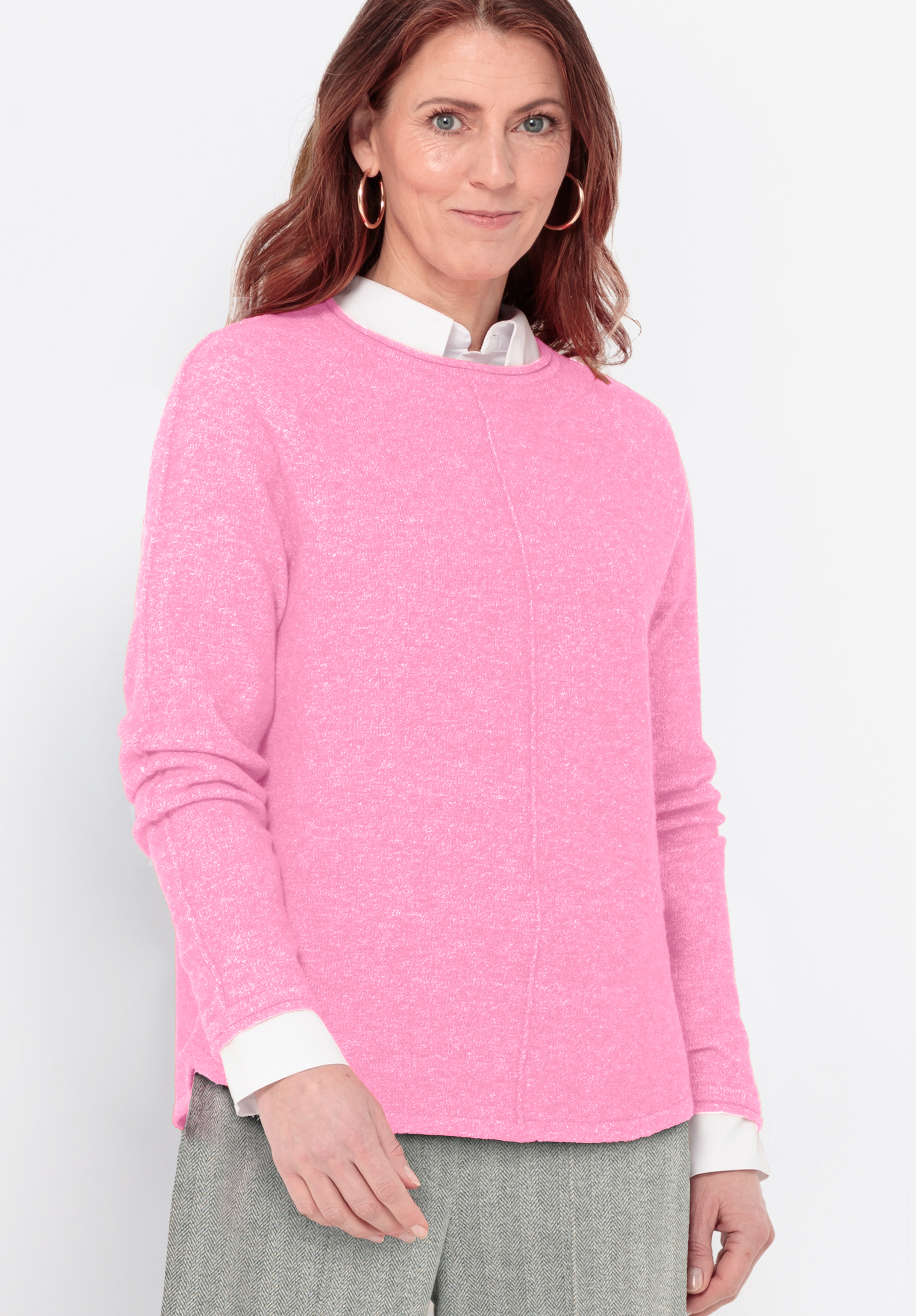 Eine Frau mit langen braunen Haaren trägt einen hellrosa Pullover über einem weißen Hemd mit Kragen und eine hellgraue Hose. Sie steht vor einem schlichten weißen Hintergrund und lächelt sanft in die Kamera.