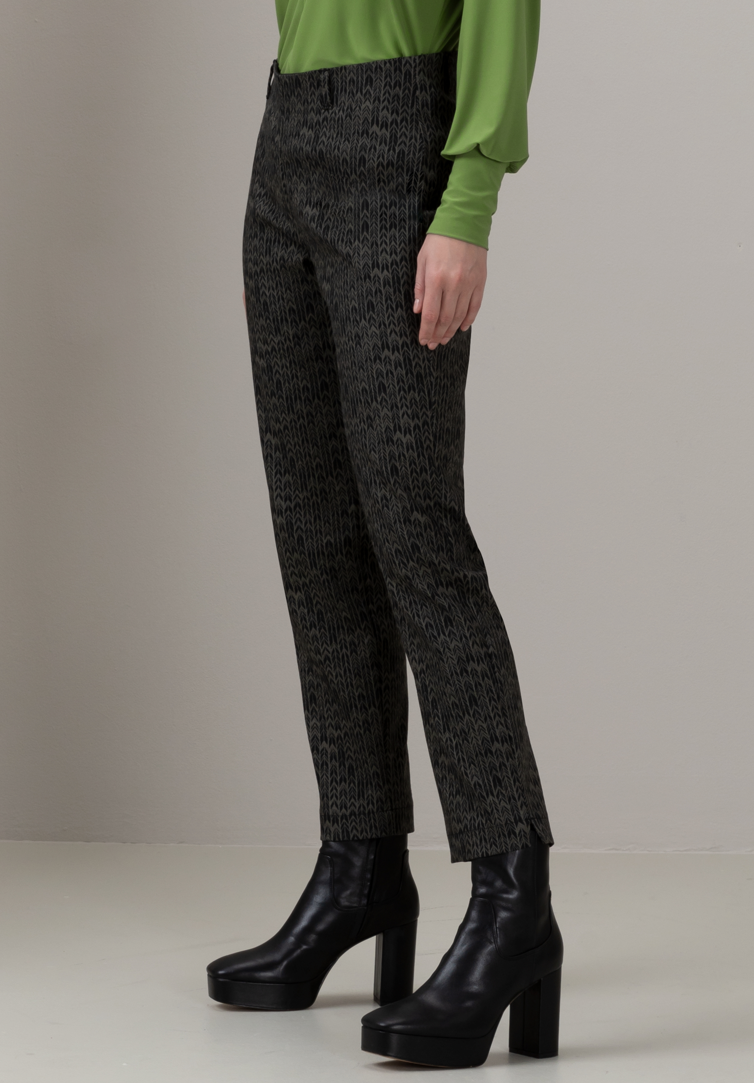 Eine Person in gemusterter dunkler Hose, leuchtend grünem Langarmshirt und schwarzen Plateaustiefeletten steht vor einem schlichten hellen Hintergrund. Nur die untere Körperhälfte ist sichtbar.