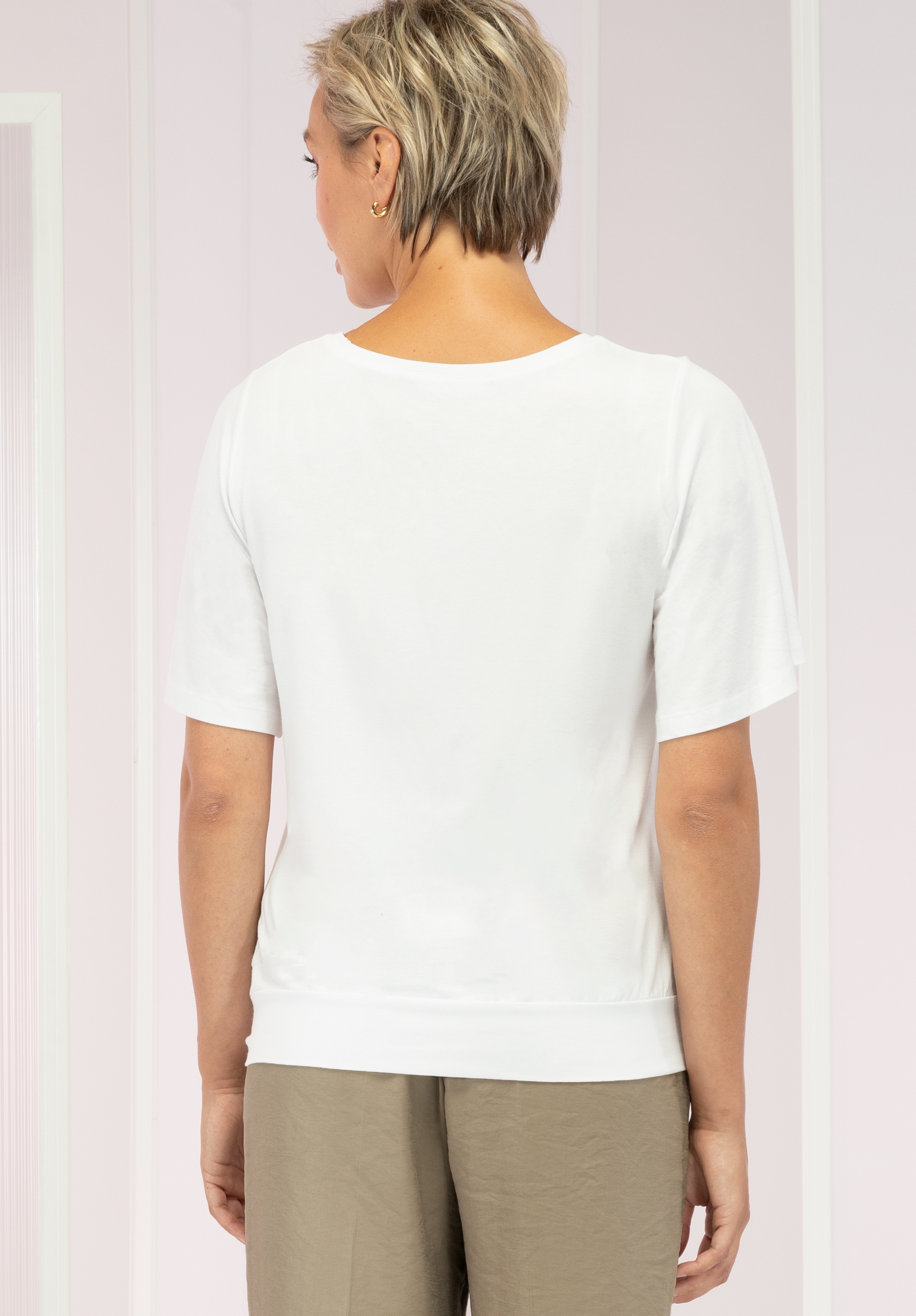 Von hinten ist eine Person mit kurzen blonden Haaren zu sehen, die ein weißes Kurzarmoberteil und hellbraune Hosen trägt und vor einem hellrosa-weiß gestreiften Hintergrund steht.