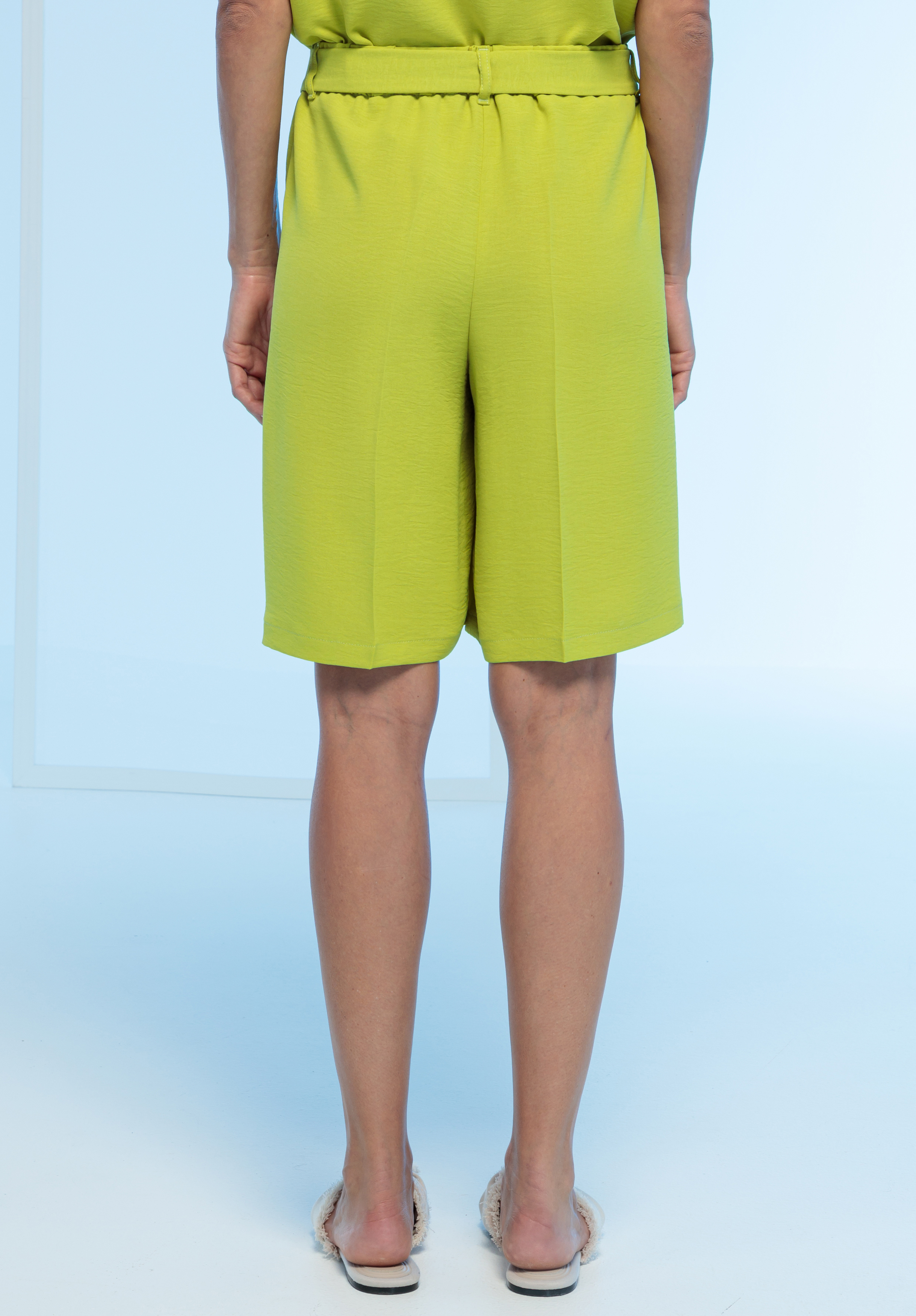 Eine Person, die auf einem hellblauen Hintergrund steht, von hinten gesehen, trägt leuchtend gelb-grüne knielange Shorts und flache Sandalen.