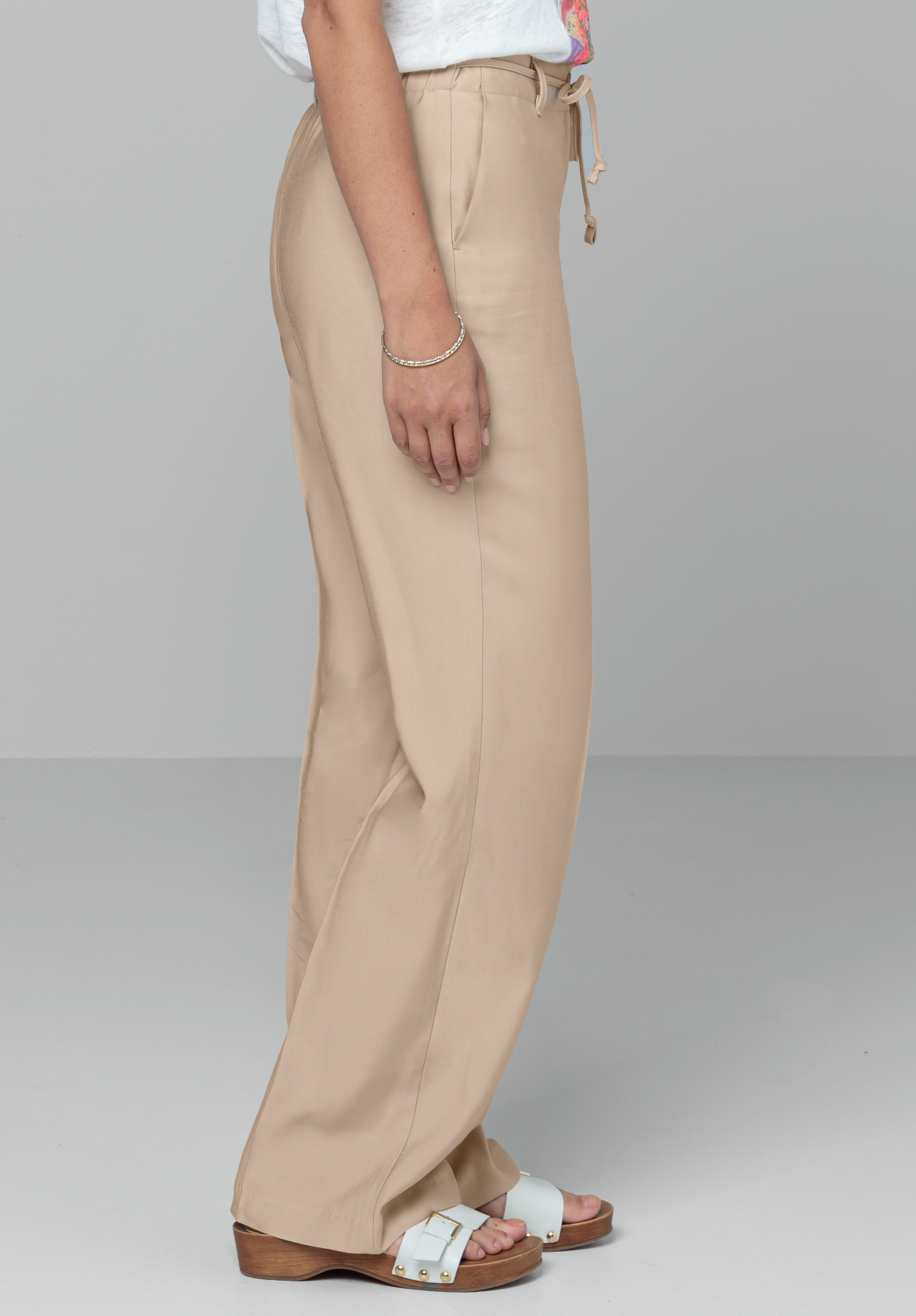 Eine seitlich stehende Person trägt beige Hosen mit weitem Bein, ein weißes Oberteil und weiße Sandalen mit Keilabsatz auf einem schlichten hellen Hintergrund.