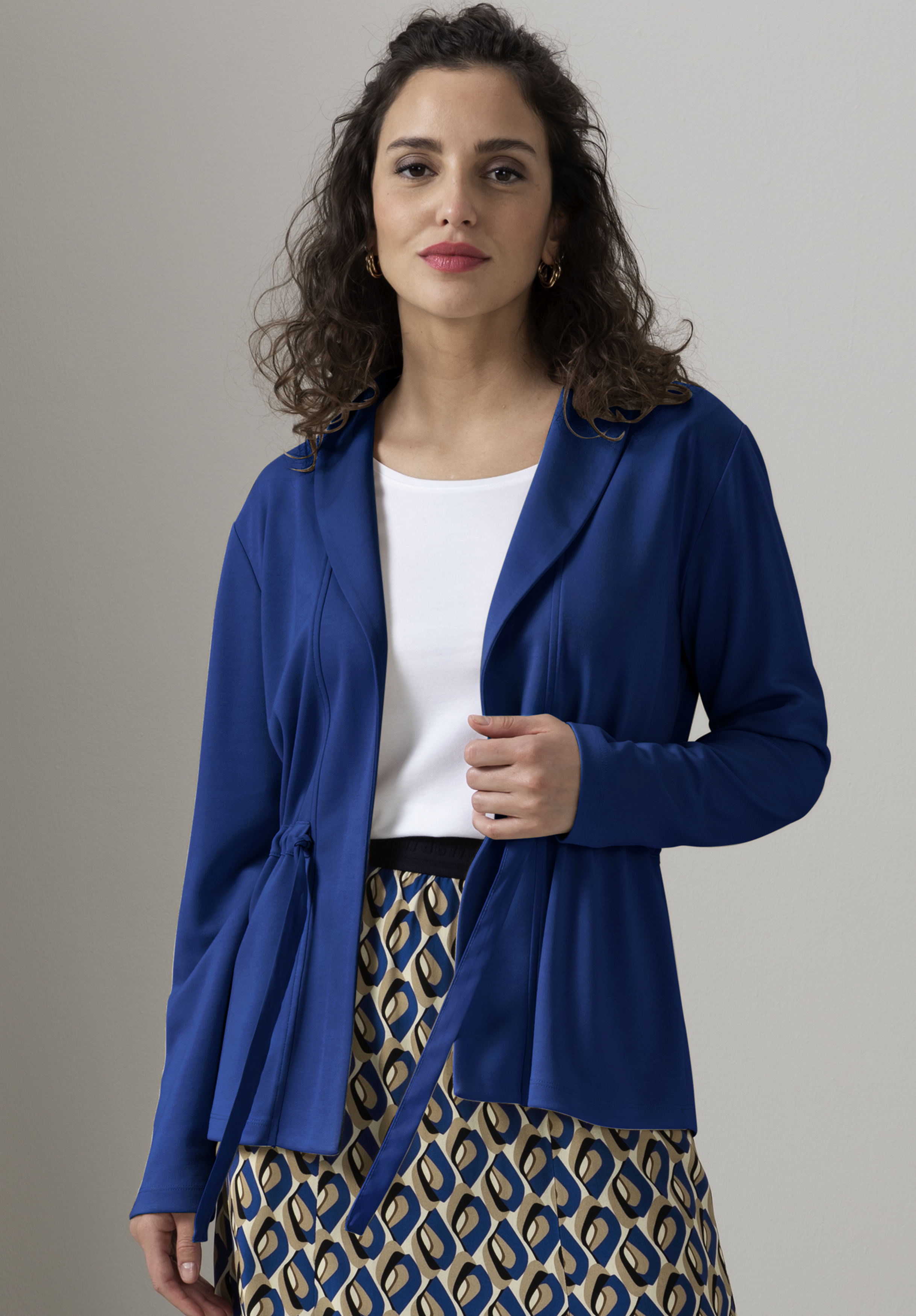 Eine Frau mit welligem braunem Haar trägt einen blauen Blazer mit Gürtel über einem weißen Oberteil und einen gemusterten Rock mit blauen, beigen und gelben Formen und steht vor einem schlichten hellen Hintergrund.