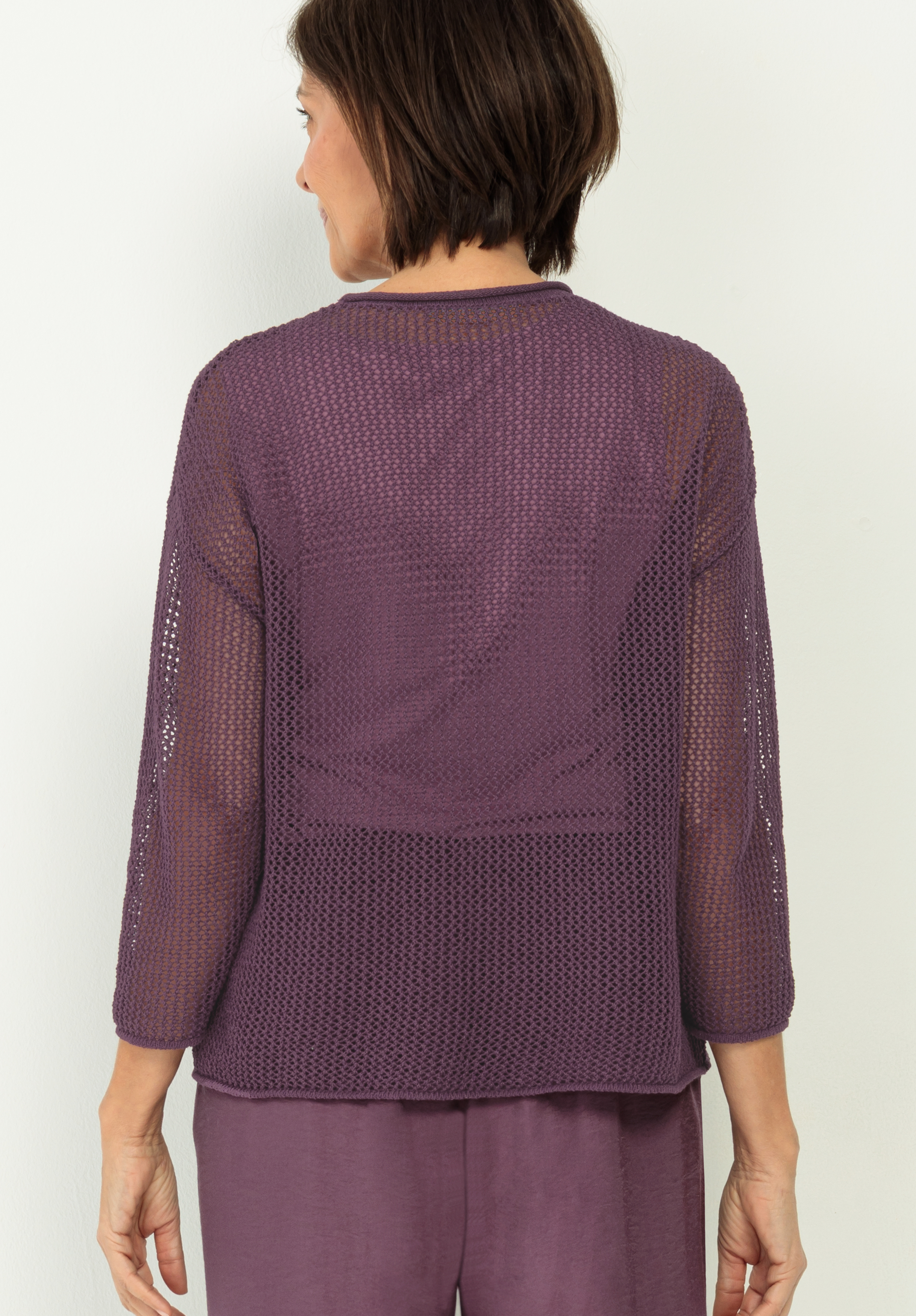 Eine Frau mit kurzen braunen Haaren wird von hinten gezeigt. Sie trägt ein durchsichtiges, strukturiertes lila Oberteil mit Dreiviertelärmeln und eine passende lila Hose. Sie steht vor einem schlichten weißen Hintergrund.