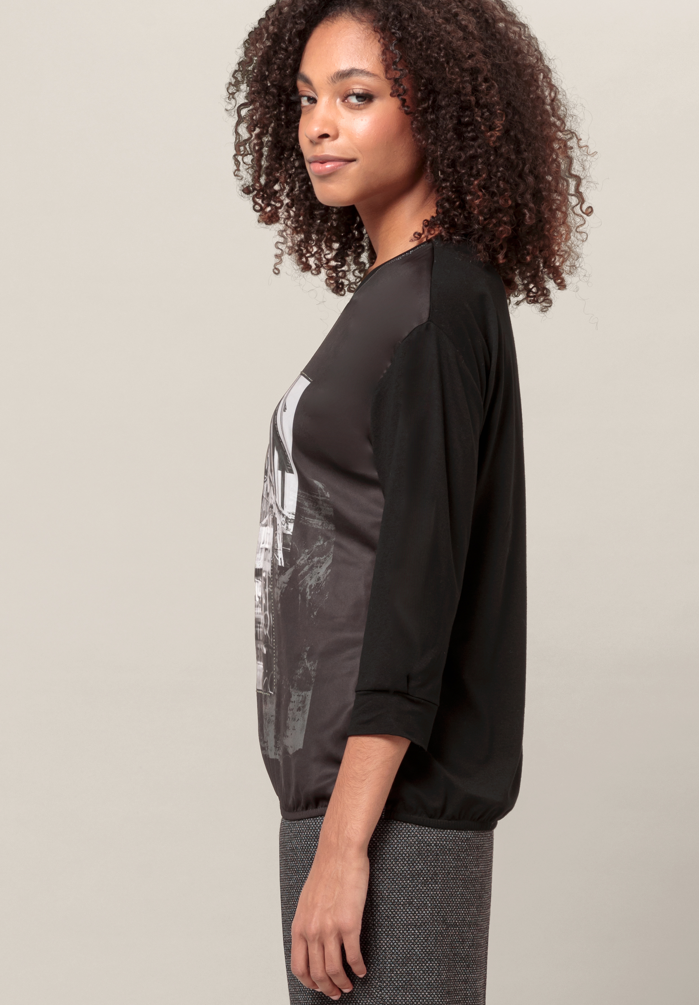 Eine Frau mit lockigem Haar steht im Profil. Sie trägt ein schwarzes Langarmshirt mit einem grafischen Muster auf der Vorderseite und einen grauen Rock vor einem schlichten hellen Hintergrund.