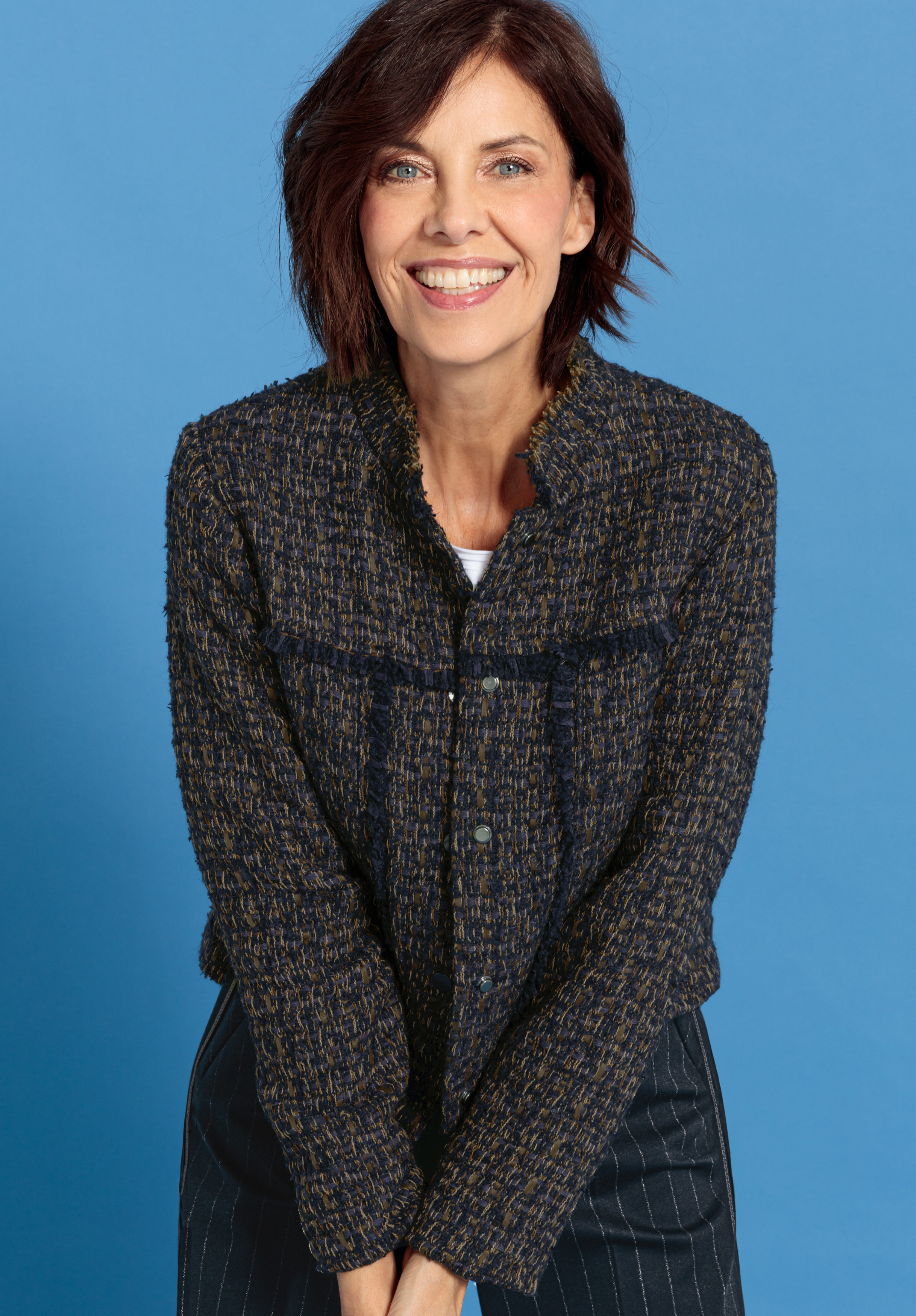 Eine Frau mit kurzen braunen Haaren lächelt in die Kamera. Sie trägt eine dunkelblaue und gelbe Jacke mit Struktur über einem weißen Oberteil und steht vor einem einfarbig blauen Hintergrund.