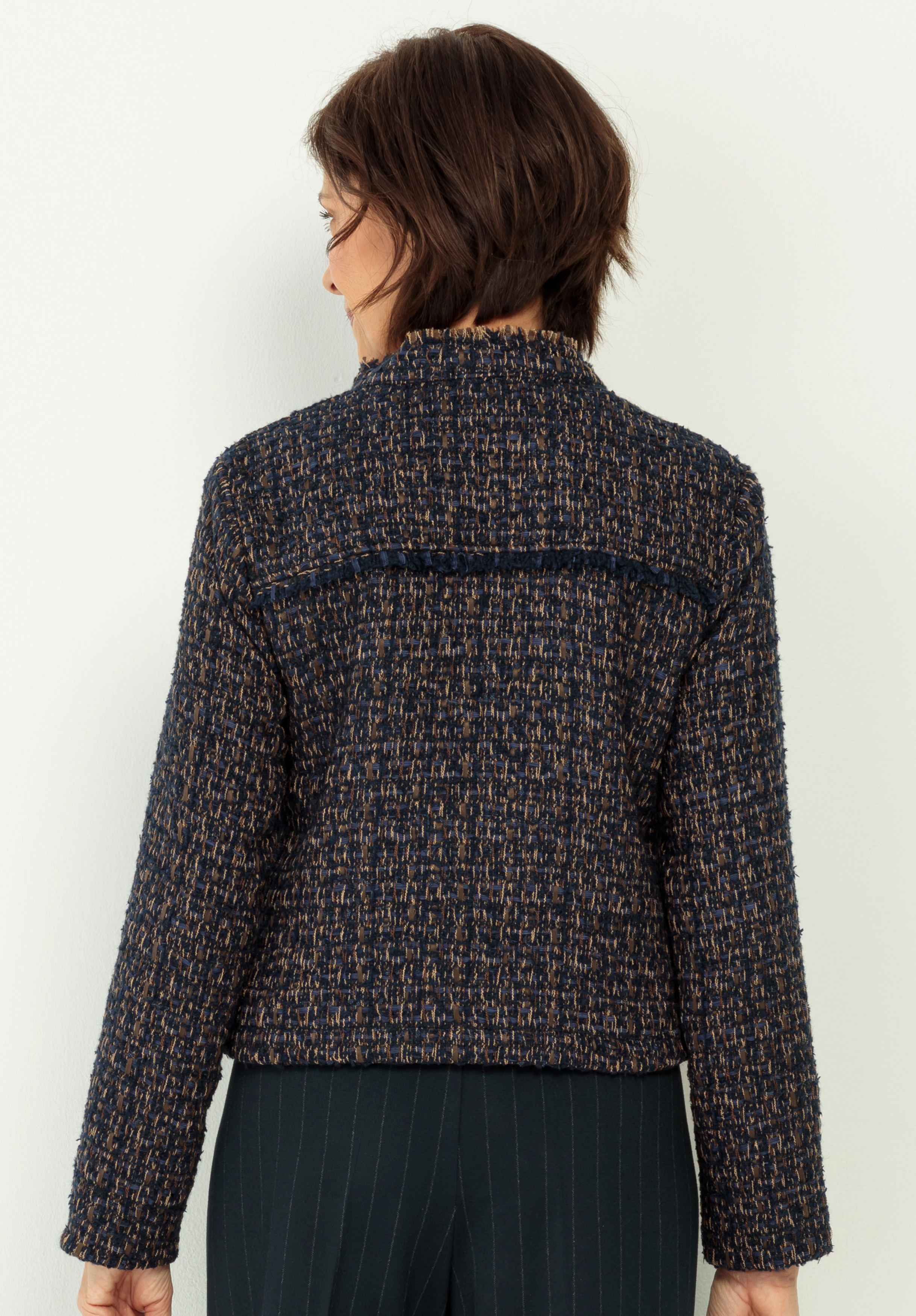 Eine Person mit kurzen braunen Haaren, von hinten gesehen, trägt eine strukturierte dunkelblaue und braune Tweedjacke mit langen Ärmeln und eine dunkle Hose. Der Hintergrund ist eine schlichte helle Wand.
