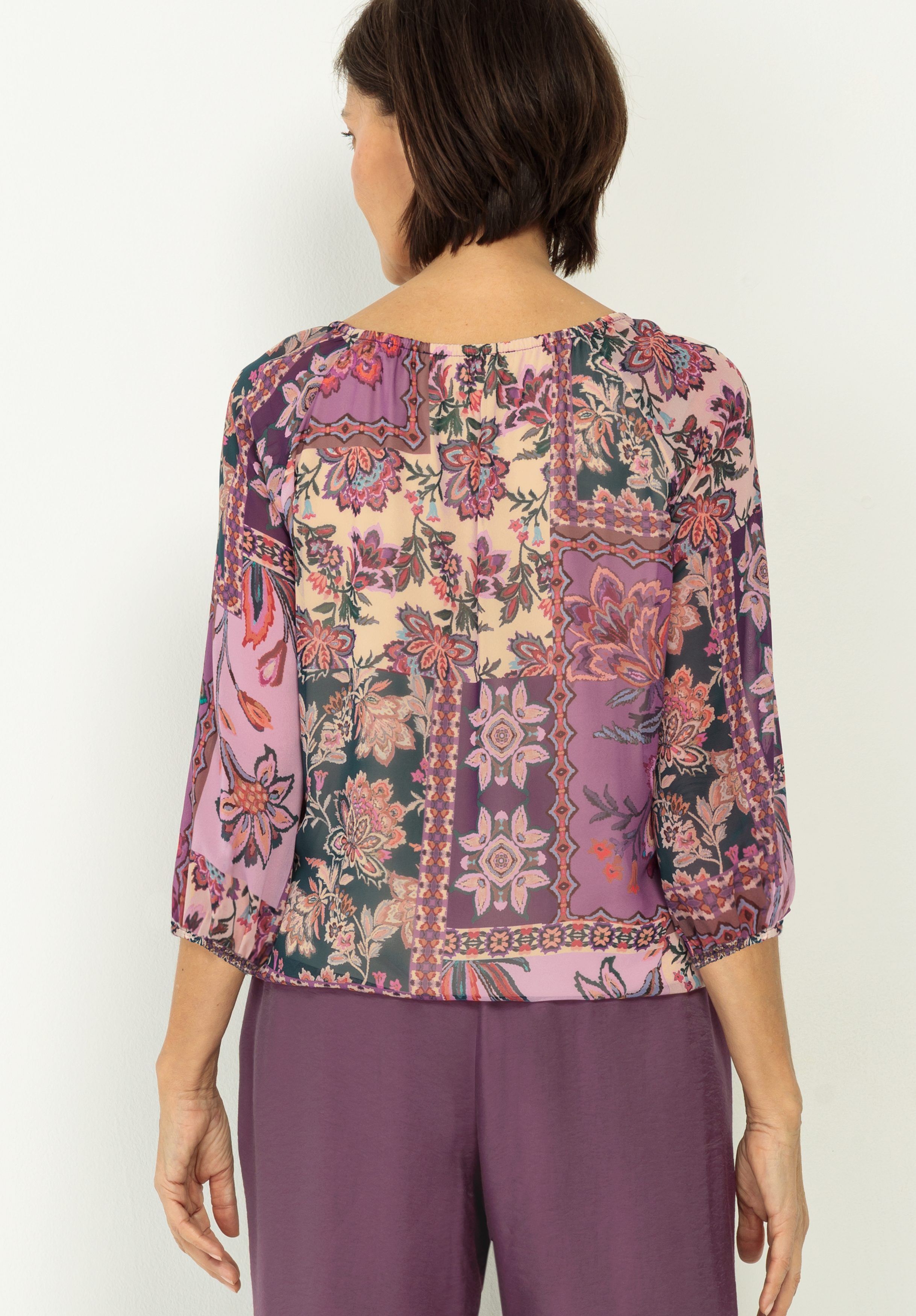 Eine Frau mit kurzen braunen Haaren ist von hinten zu sehen. Sie trägt eine farbenfrohe, blumengemusterte Bluse mit lila, rosa und grünen Farbtönen und eine locker sitzende lila Hose.