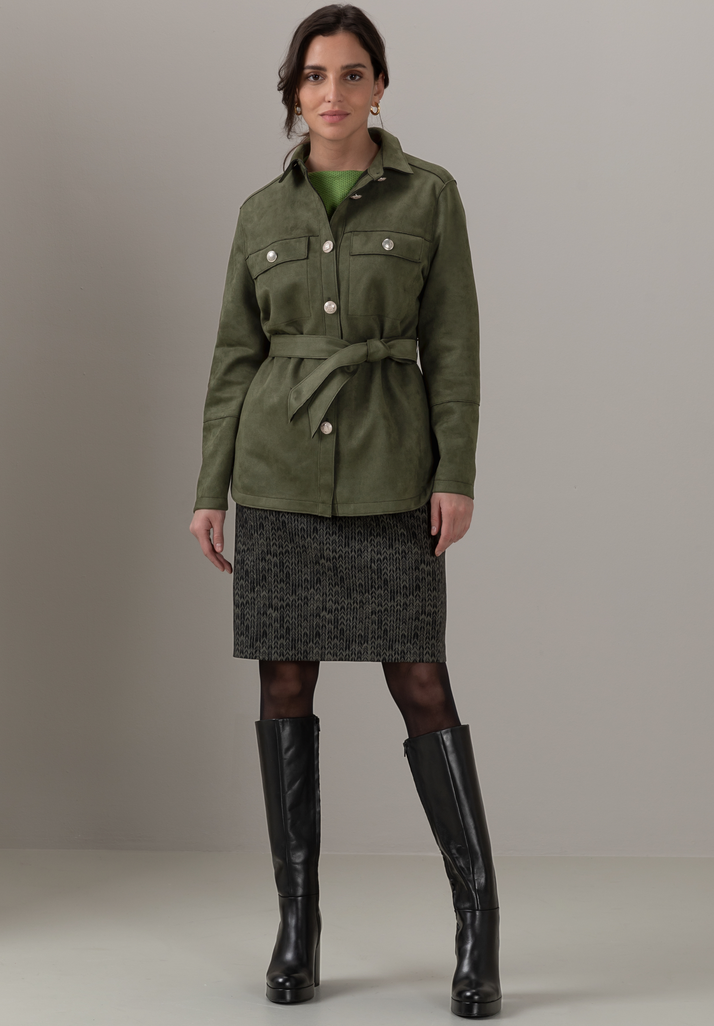 Eine Frau steht vor einem schlichten Hintergrund. Sie trägt eine olivgrüne Jacke mit Gürtel, einen gemusterten knielangen Rock, schwarze Strumpfhosen und kniehohe schwarze Stiefel. Sie hat dunkles, nach hinten frisiertes Haar und blickt in die Kamera.
