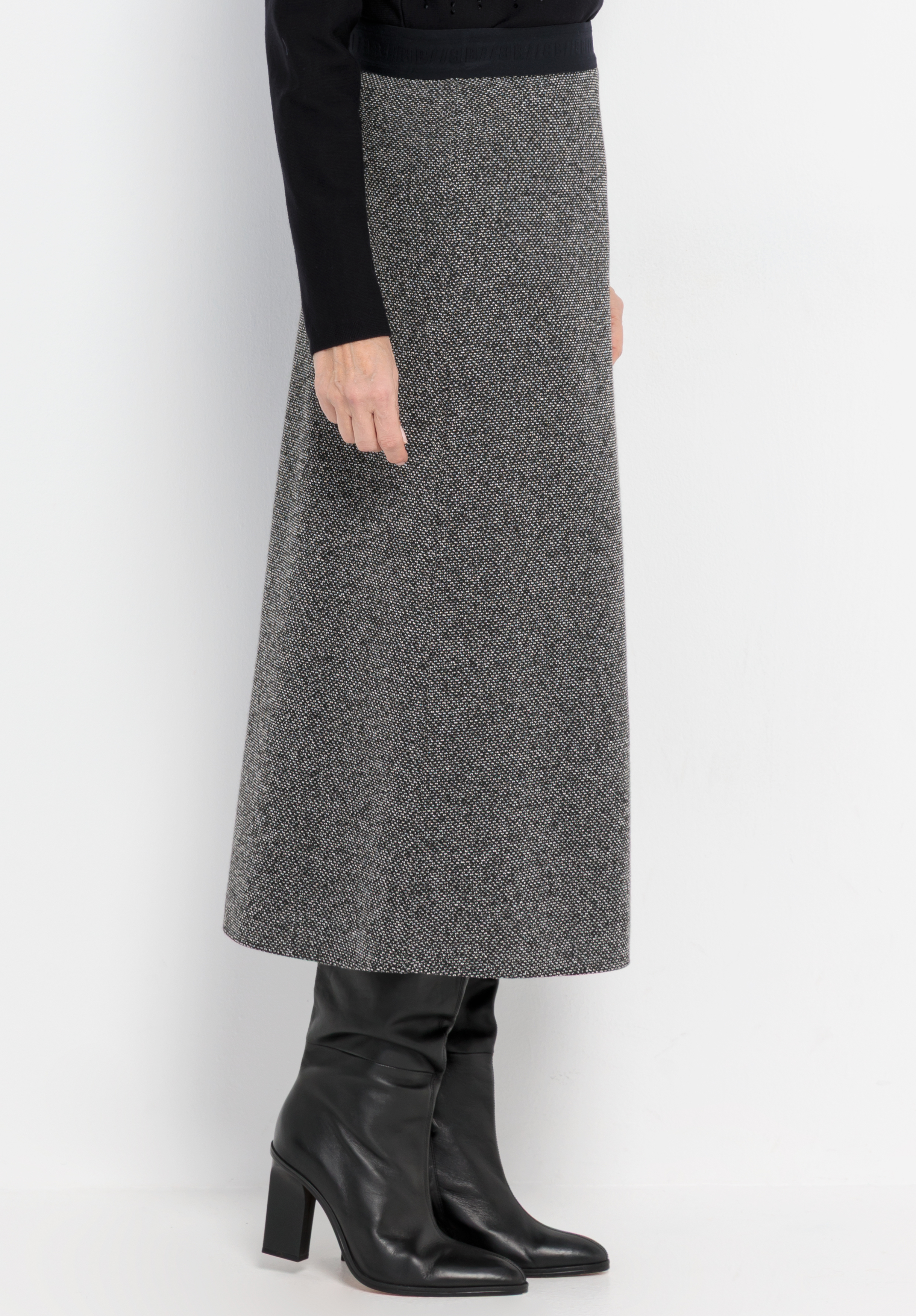 Eine Person, die einen langen, grauen Rock im Fischgrätenmuster trägt, dazu schwarze kniehohe Stiefel mit Absatz und ein schwarzes langärmeliges Oberteil, vor einem schlichten weißen Hintergrund stehend.
