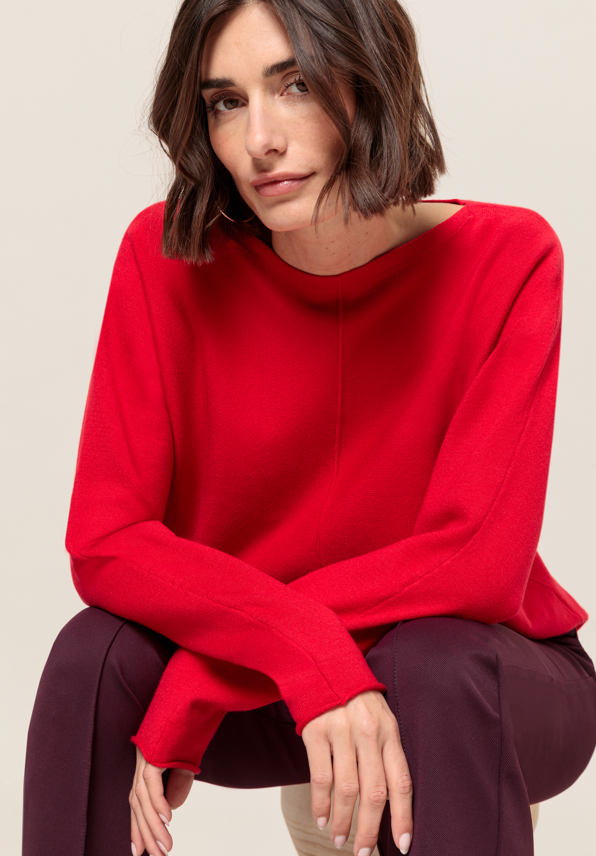 Eine Frau mit kinnlangem braunem Haar, die ein leuchtend rotes Langarmoberteil und eine dunkle Hose trägt, sitzt leicht nach vorne gebeugt da und blickt mit einem subtilen Lächeln vor einem hellen, neutralen Hintergrund in die Kamera.
