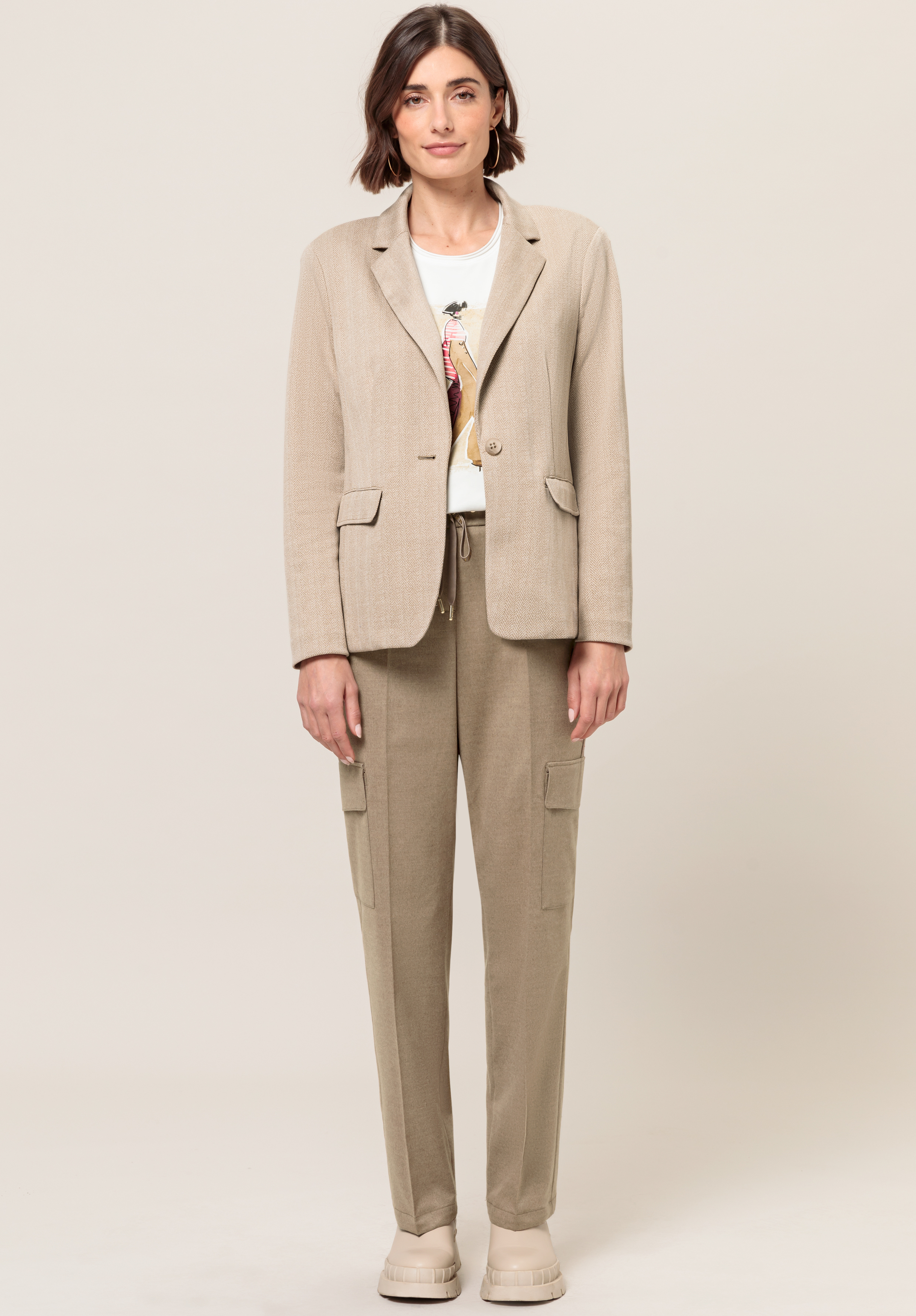 Eine Frau mit kurzen braunen Haaren trägt einen beigen Blazer über einem weißen bedruckten T-Shirt, beige Cargohosen und beige Schuhe und steht vor einem schlichten hellen Hintergrund.
