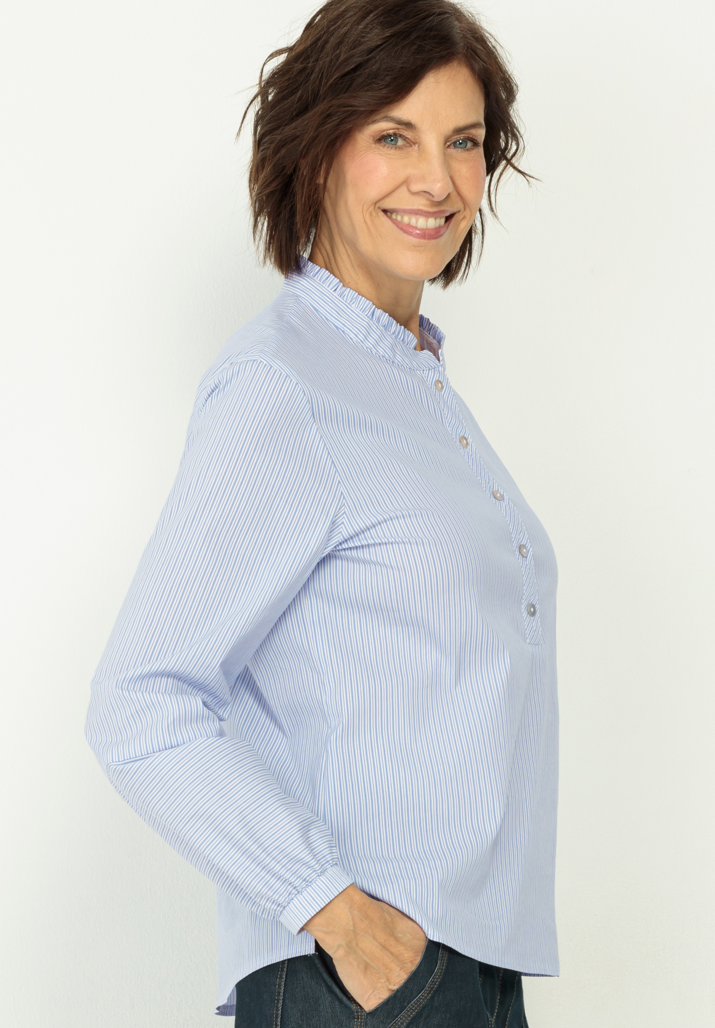 Eine Frau mit kurzen braunen Haaren, die ein hellblau gestreiftes Button-up-Hemd und dunkle Jeans trägt, steht seitlich mit einer Hand in der Tasche und lächelt in die Kamera vor einem schlichten hellen Hintergrund.