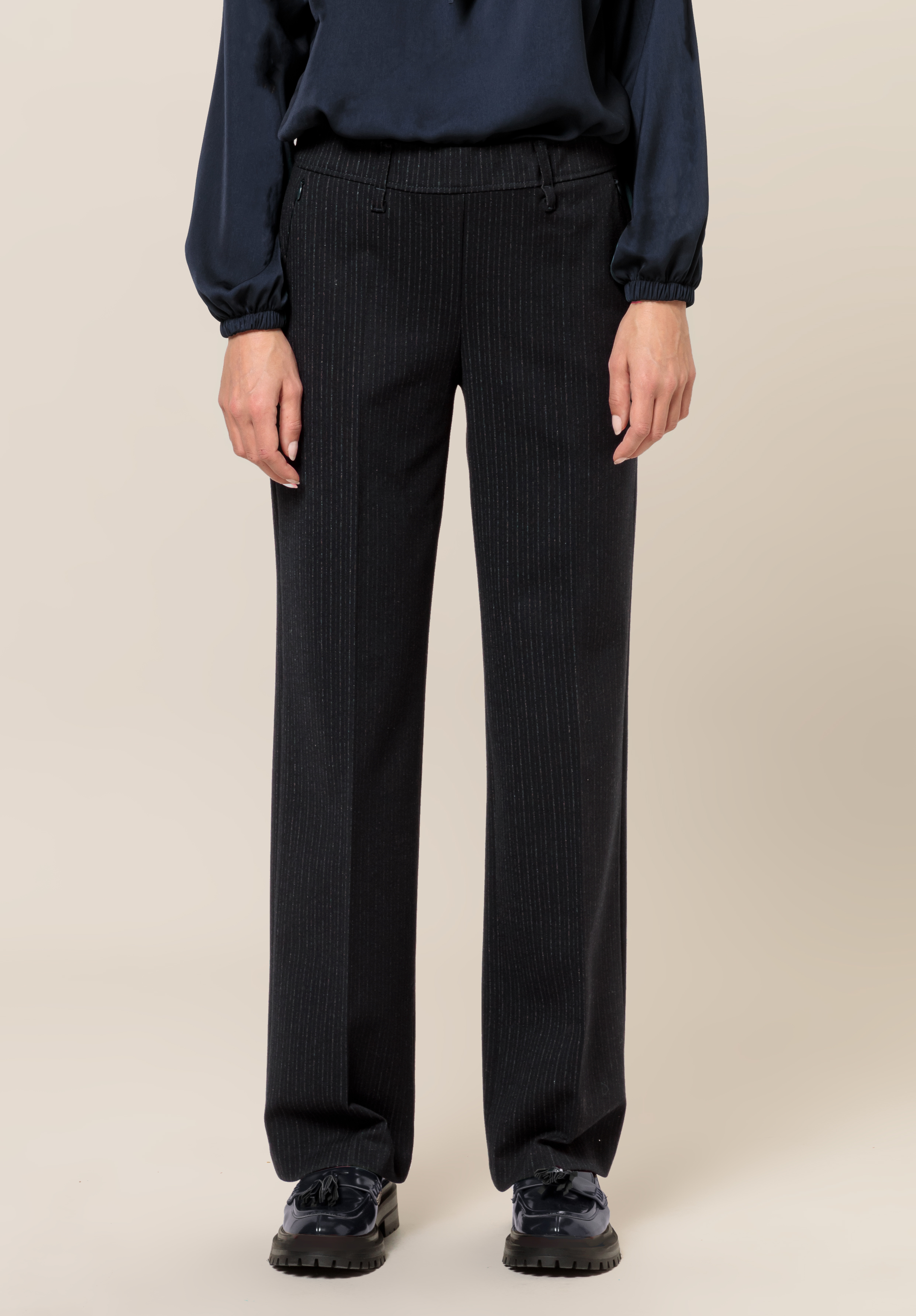 Eine Person, die ein dunkelblaues Langarmoberteil, eine schwarze Hose mit weitem Bein und Nadelstreifen sowie schwarze klobige Schuhe trägt, steht vor einem schlichten beigen Hintergrund.