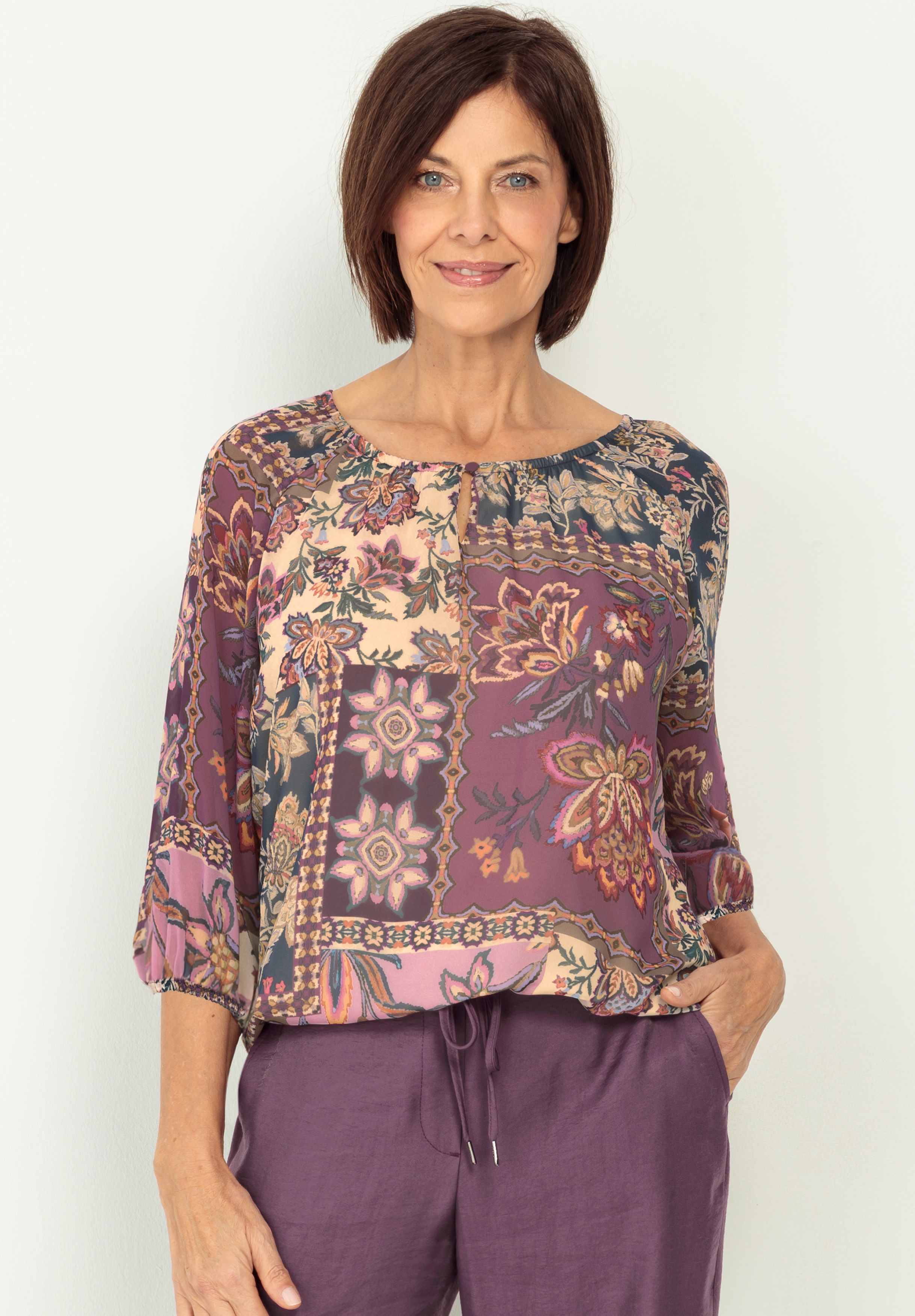Eine Frau mit kurzen braunen Haaren trägt eine lockere, gemusterte Bluse in lila und cremefarbenen Tönen mit Blumenmustern, dazu eine lila Hose. Sie lächelt und steht vor einem schlichten hellen Hintergrund.