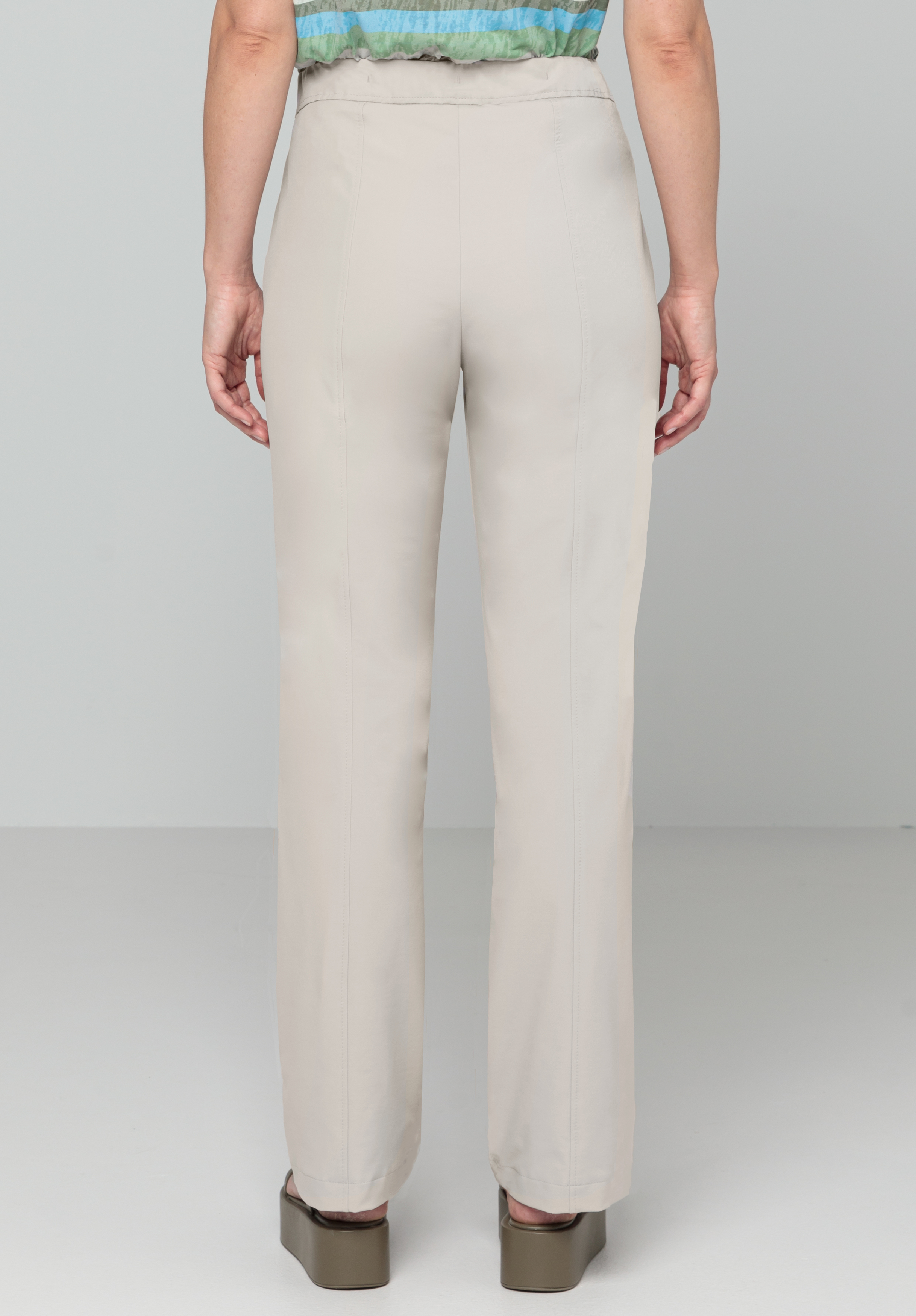 Eine Person steht mit dem Rücken zur Kamera und trägt hellbeige Hosen mit geradem Bein, ein grün-blau gemustertes Oberteil und braune Plateauschuhe vor einem schlichten hellgrauen Hintergrund.