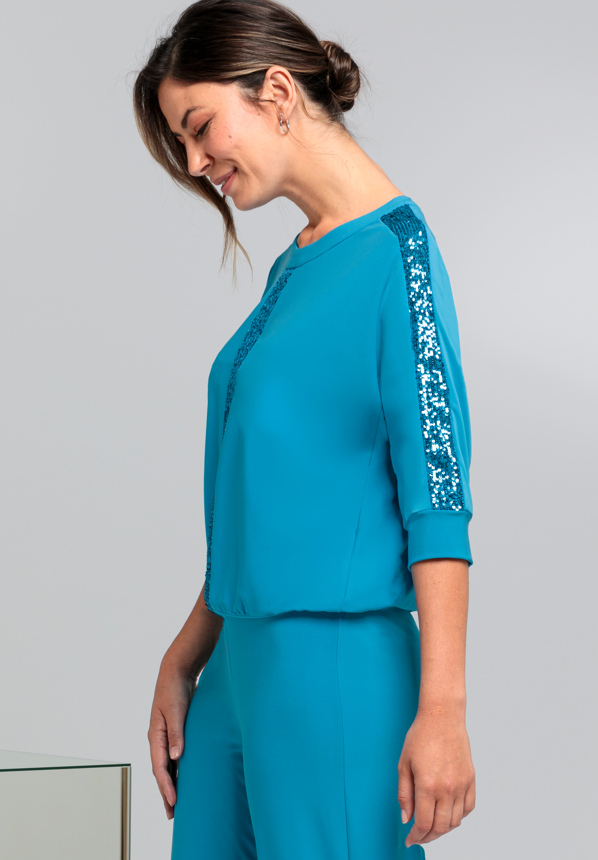 Eine Frau in einem blaugrünen Outfit mit Paillettenstreifen an Ärmeln und Vorderseite steht im Profil, lächelt sanft und hat den Kopf leicht nach unten geneigt. Ihr Haar ist zurückgebunden und sie trägt kleine Creolen.