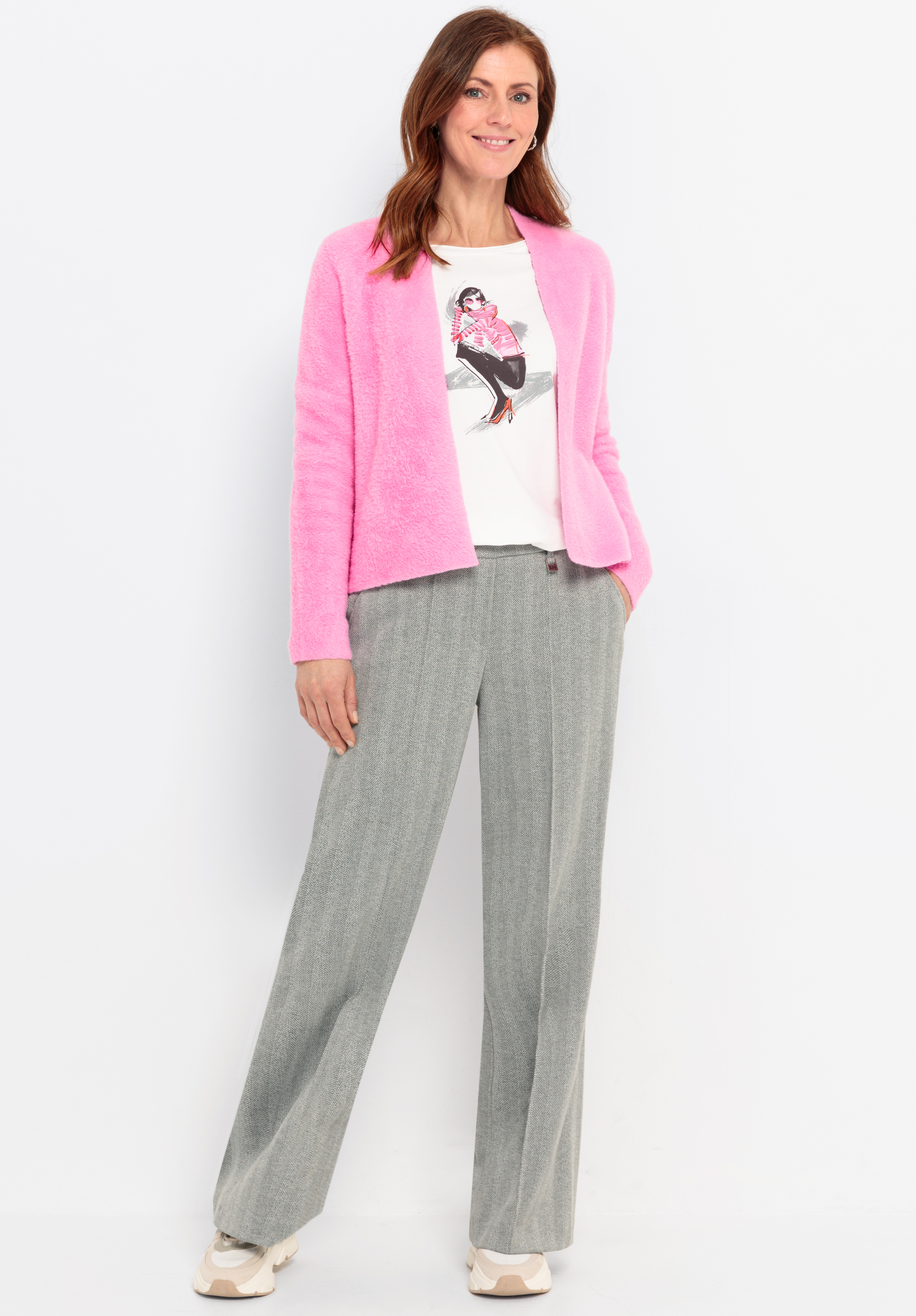 Eine Frau steht lächelnd vor einem weißen Hintergrund und trägt eine leuchtend rosa Strickjacke, ein weißes grafisches T-Shirt, eine graue Hose mit weitem Bein und beigefarbene Turnschuhe. Ihre linke Hand steckt in ihrer Tasche.