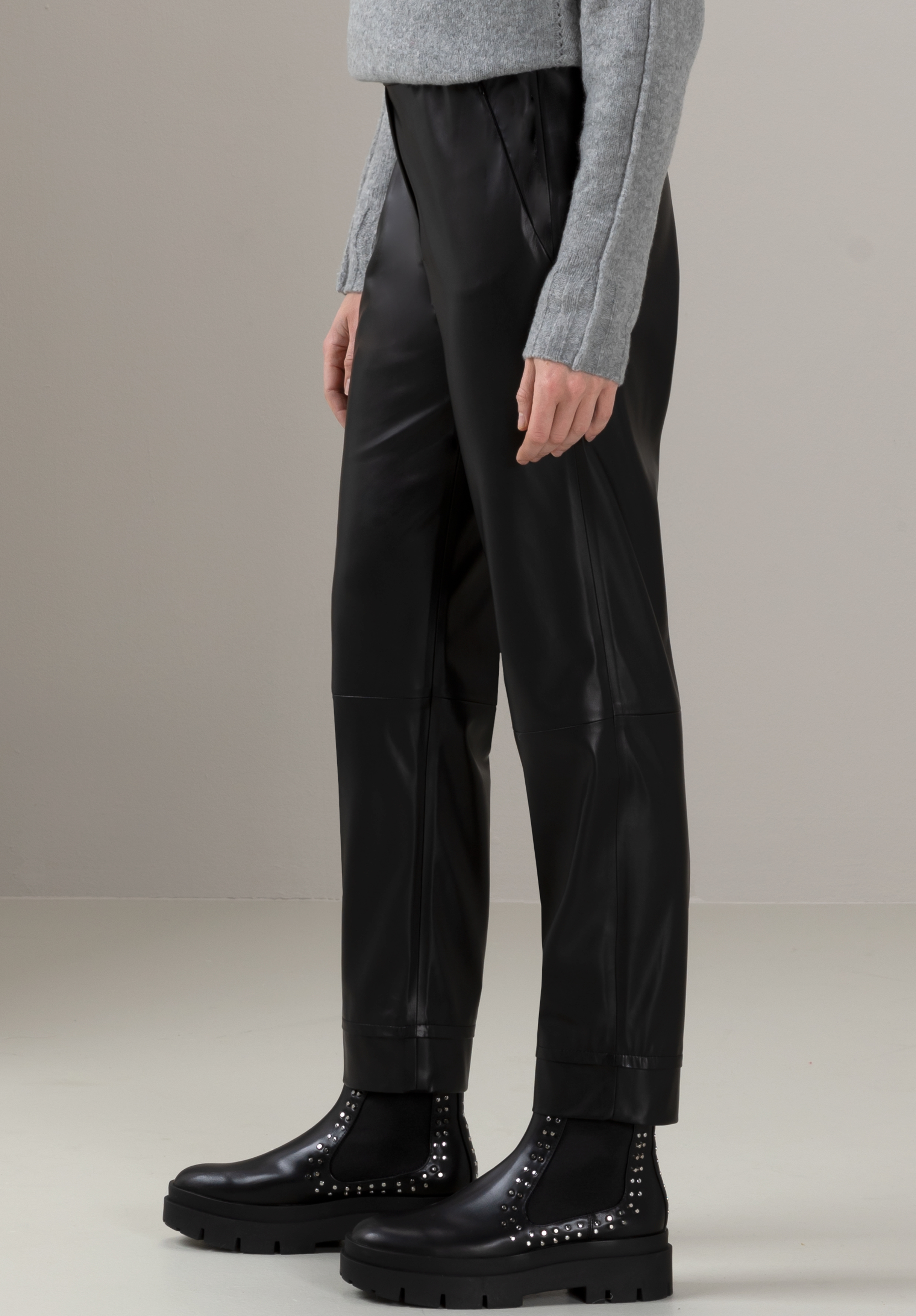 Eine Person in einem grauen Pullover, einer schwarzen Kunstlederhose und schwarzen Stiefeletten mit silbernen Nieten steht vor einem schlichten, hellen Hintergrund. Das Bild ist unterhalb der Schultern abgeschnitten.
