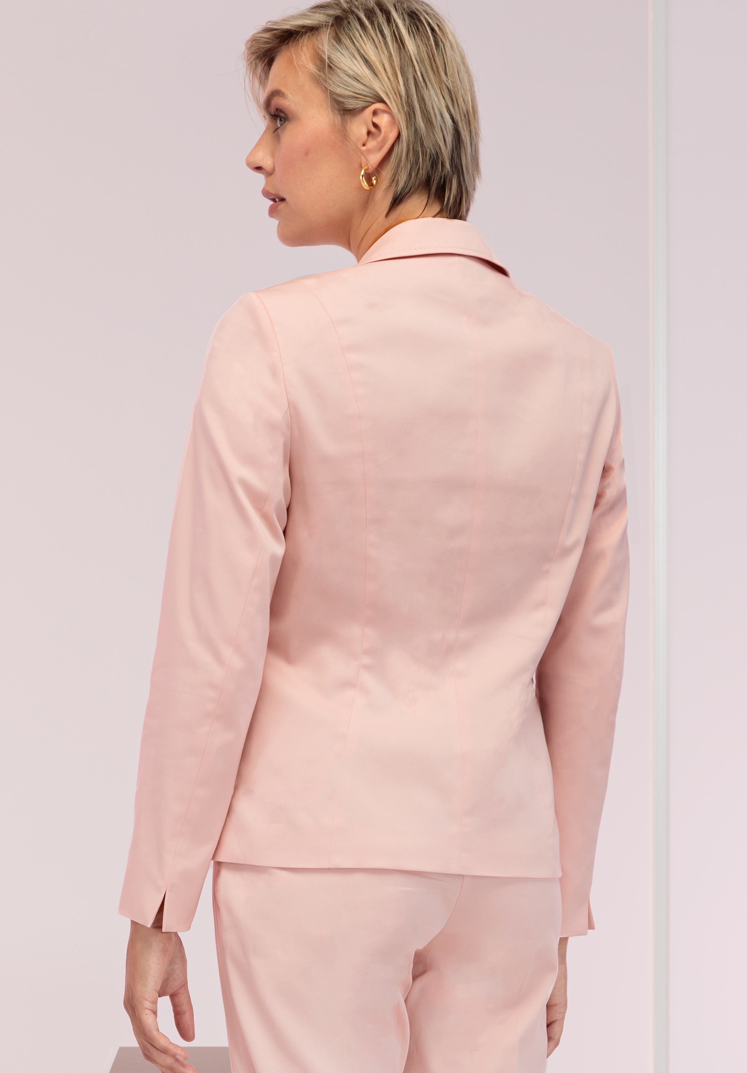Eine Person mit kurzen blonden Haaren, einem hellrosa Blazer und der dazu passenden Hose steht mit abgewandtem Blick da und zeigt die Rückseite des Outfits. Der Hintergrund ist in einem sanften Hellrosa gehalten.