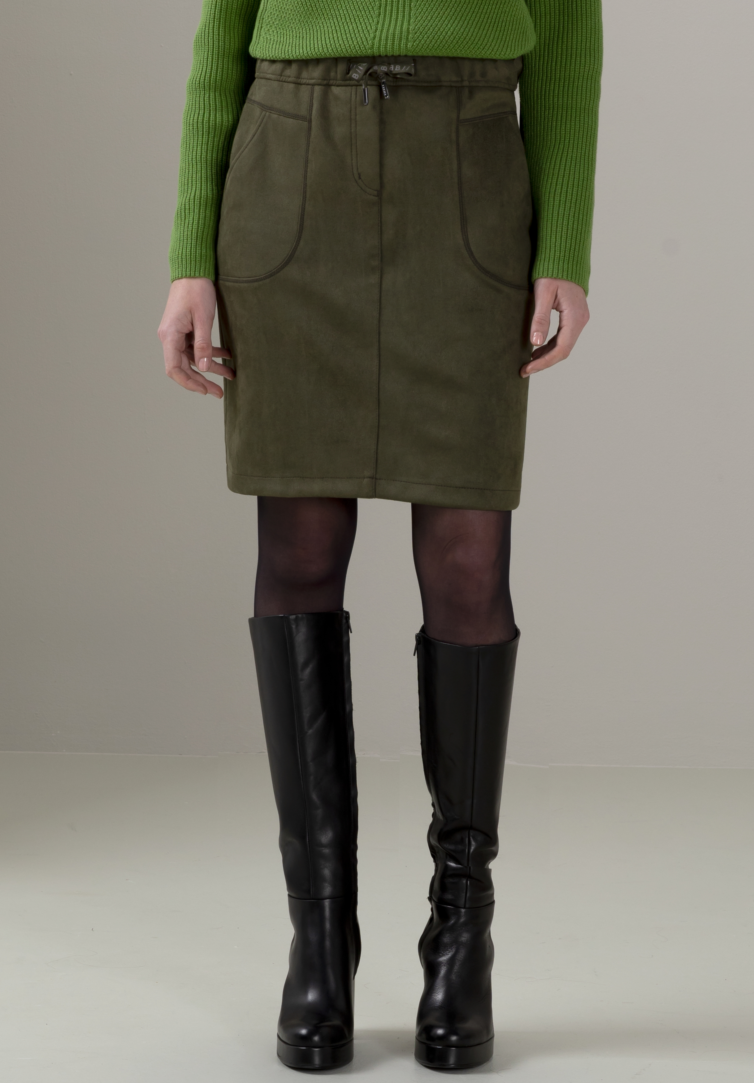 Schlupfrock KINI Eine Person, die einen grünen Langarmpullover, einen olivgrünen Rock mit Vordertaschen und Kordelzug, schwarze Strumpfhosen und kniehohe schwarze Stiefel trägt, steht vor einem schlichten hellen Hintergrund.