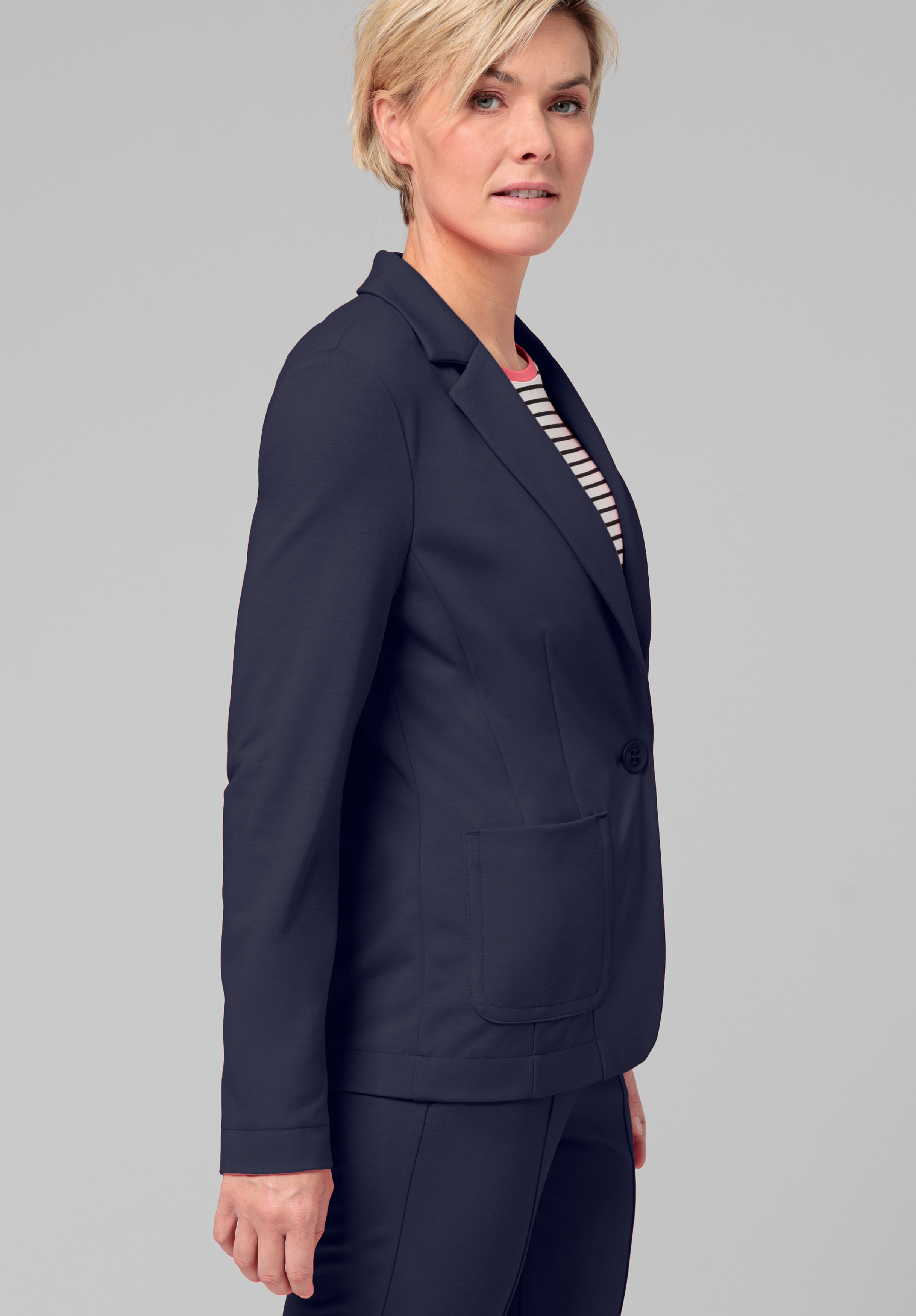 Eine Frau mit kurzen blonden Haaren trägt einen dunklen Blazer und eine passende Hose über einem gestreiften Hemd, steht seitlich und blickt mit neutralem Gesichtsausdruck vor einem schlichten grauen Hintergrund in die Kamera.