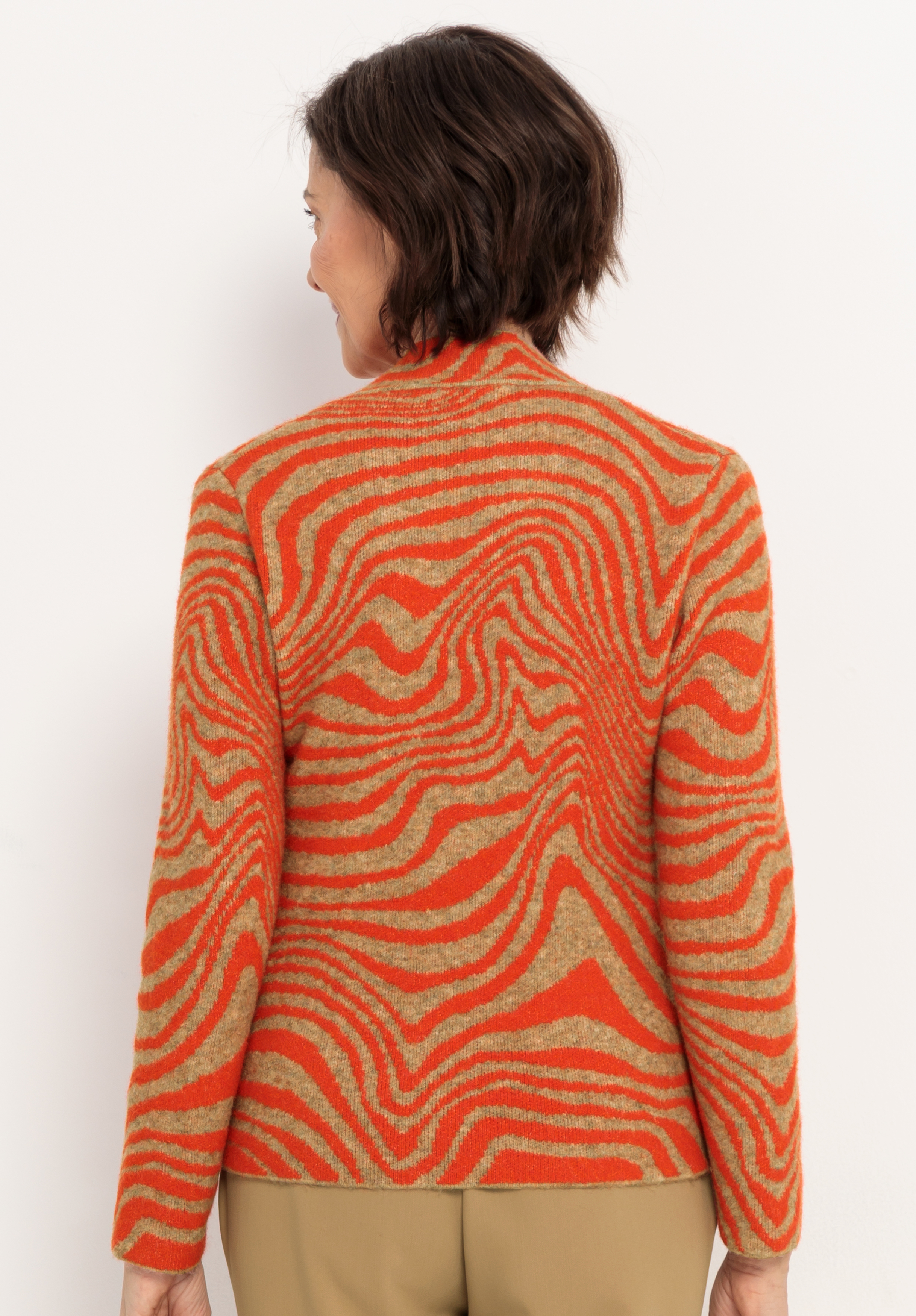 Eine Person mit kurzen braunen Haaren, von hinten gesehen, trägt einen langärmeligen Pullover mit einem orangefarbenen, gewellten Zebrastreifenmuster und eine hellbraune Hose und steht vor einem schlichten weißen Hintergrund.