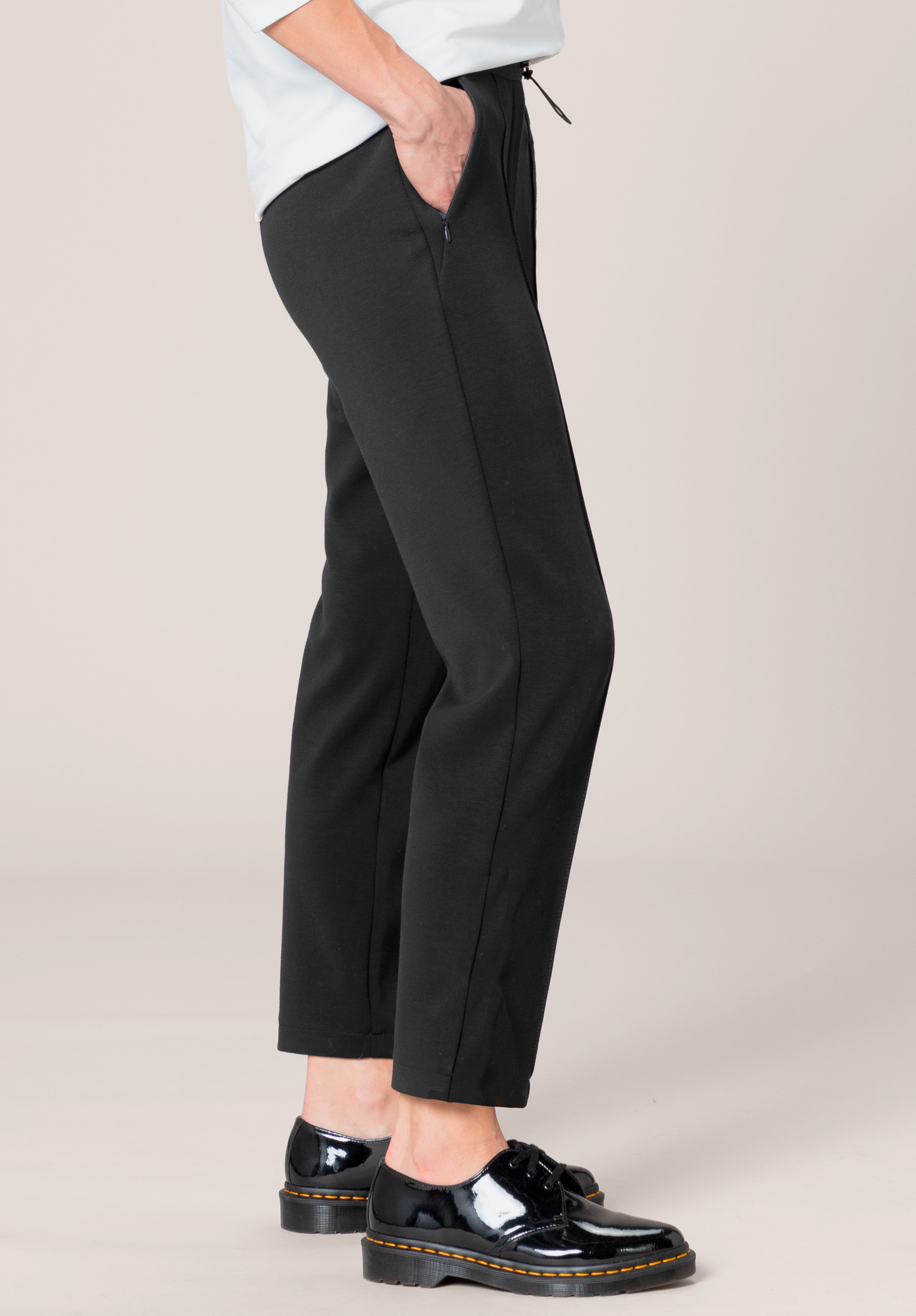 Eine Person steht seitlich, trägt eine schwarze Hose mit geradem Bein und glänzende schwarze Schnürschuhe und hat eine Hand in der Tasche. Der Hintergrund ist schlicht und hell.
