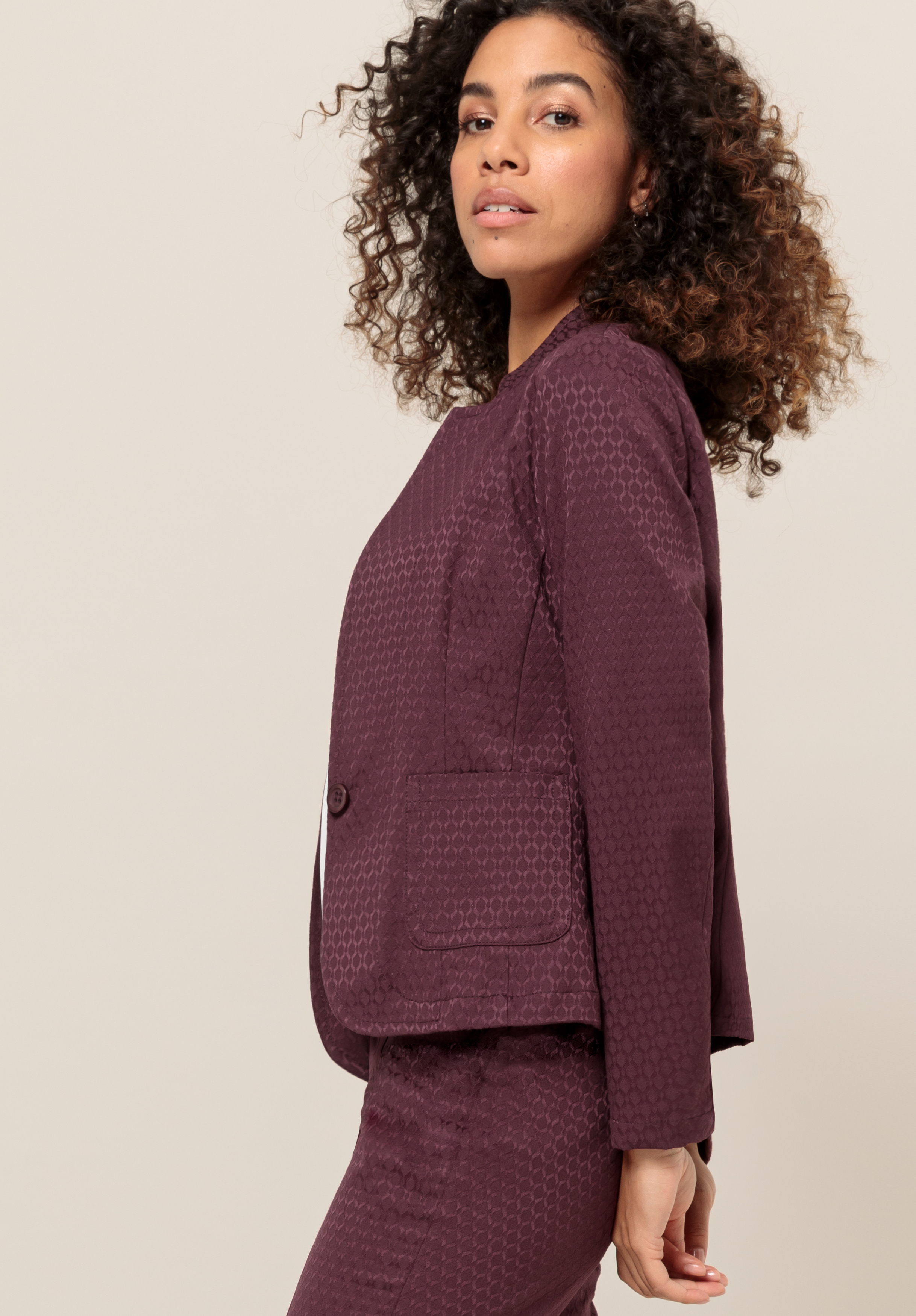 Eine Frau mit lockigem Haar, die einen strukturierten, dunkelvioletten Blazer und eine passende Hose trägt, steht im Profil vor einem neutralen Hintergrund und blickt selbstbewusst in die Kamera.