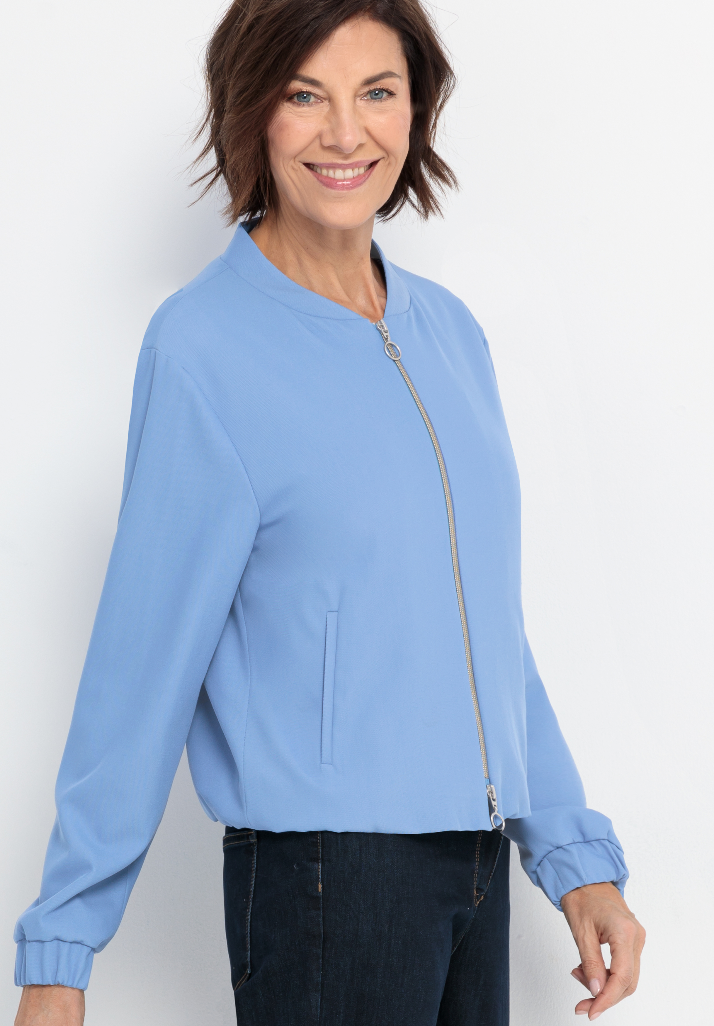 Eine Frau mit kurzen braunen Haaren lächelt, während sie eine hellblaue Jacke mit Reißverschluss und dunkle Jeans trägt und vor einem schlichten weißen Hintergrund steht.