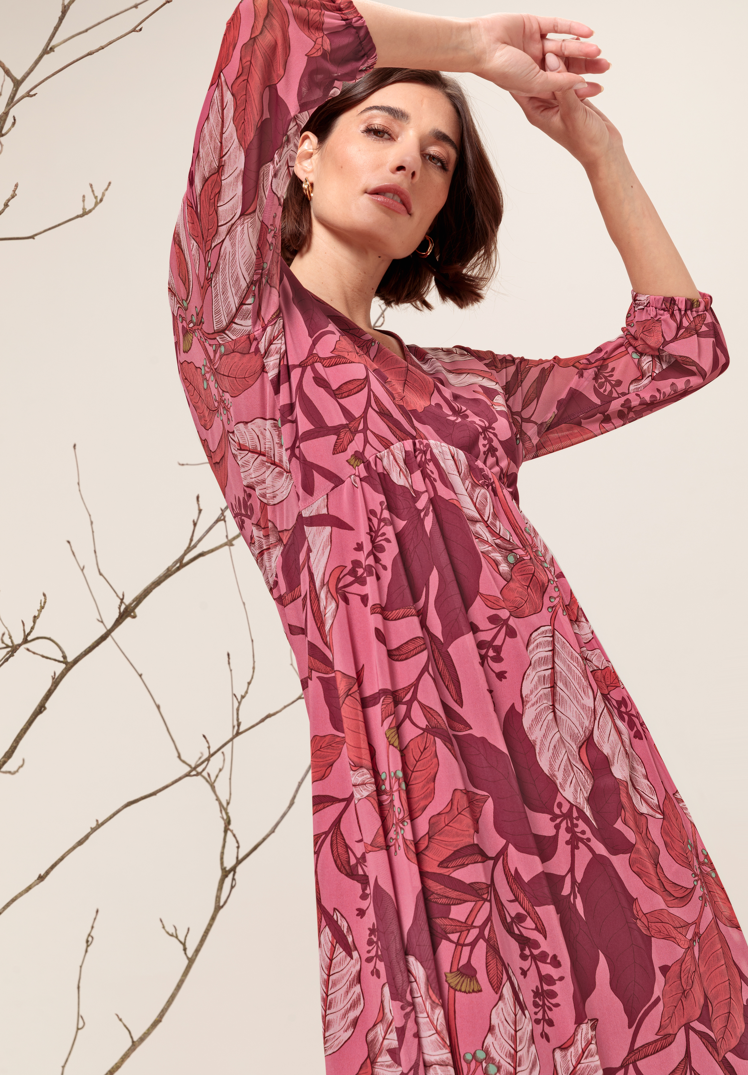 Eine Frau in einem rosa-roten Blumenkleid posiert mit über dem Kopf erhobenen Armen. Sie steht neben kahlen, dünnen Ästen vor einem hellen Hintergrund.
