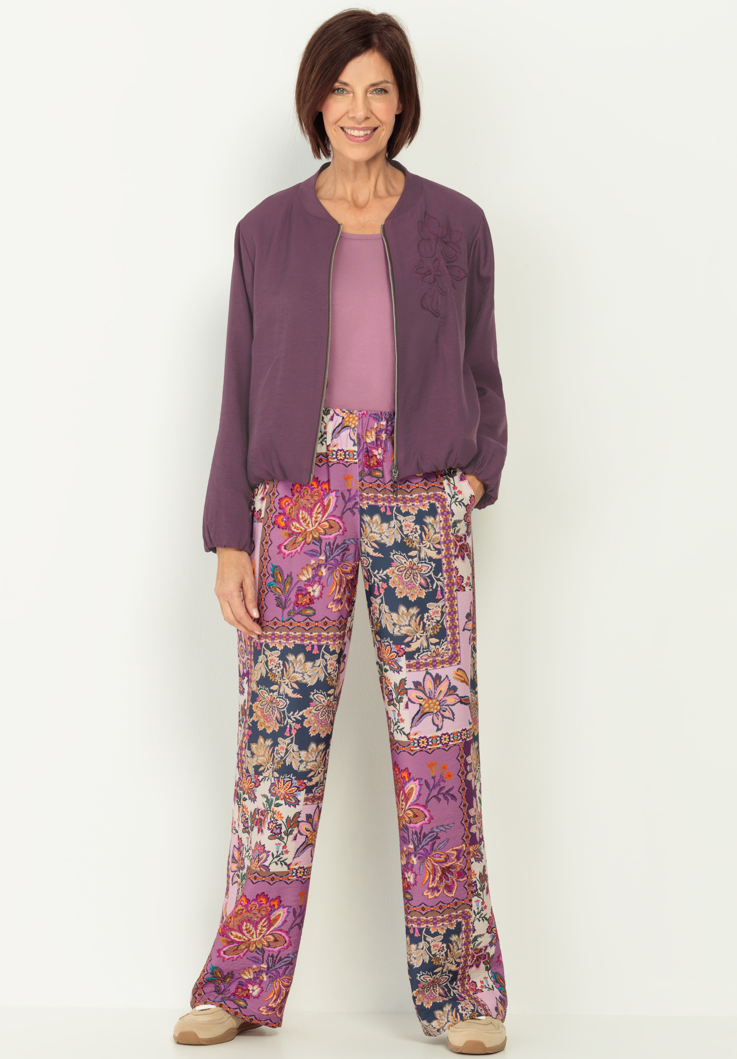 Eine Frau steht lächelnd da und trägt eine lila Jacke, ein dazu passendes Oberteil und eine weitbeinige, geblümte Patchwork-Hose in Rosa-, Lila-, Blau- und Beigetönen. Sie hat kurzes braunes Haar und steht vor einem schlichten weißen Hintergrund.