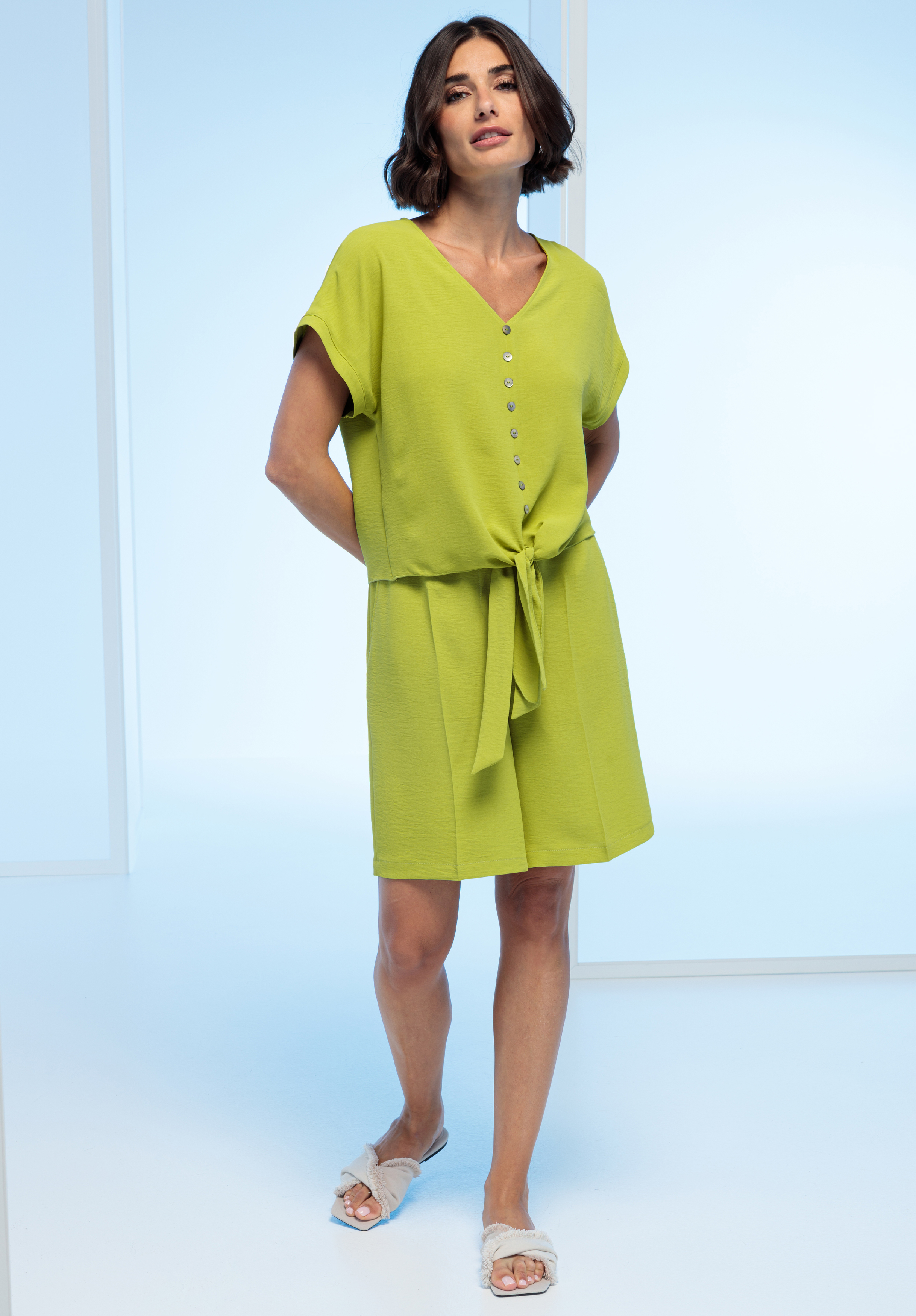 Eine Frau trägt ein lindgrünes, kurzärmeliges Kleid mit Knöpfen vorne und einer Krawatte in der Taille, gepaart mit weißen Slide-Sandalen.