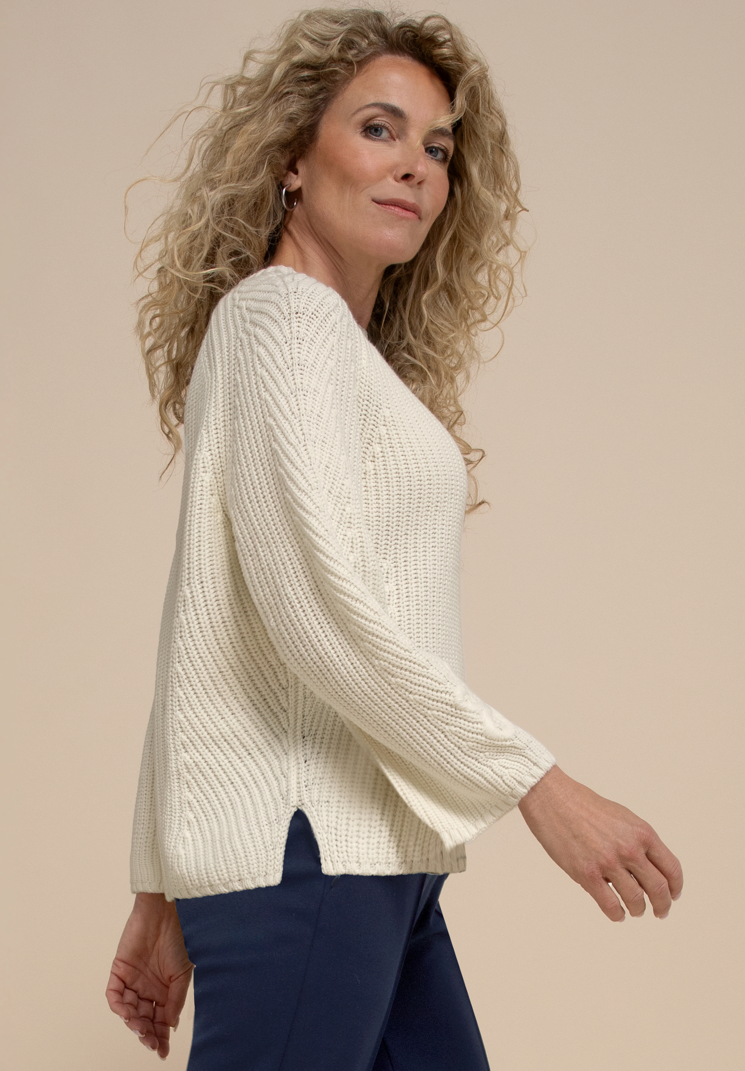 Eine Frau mit langen, lockigen blonden Haaren, die einen weißen Strickpullover und eine marineblaue Hose trägt, geht selbstbewusst vor einem beigen Hintergrund und blickt dabei leicht über ihre Schulter.