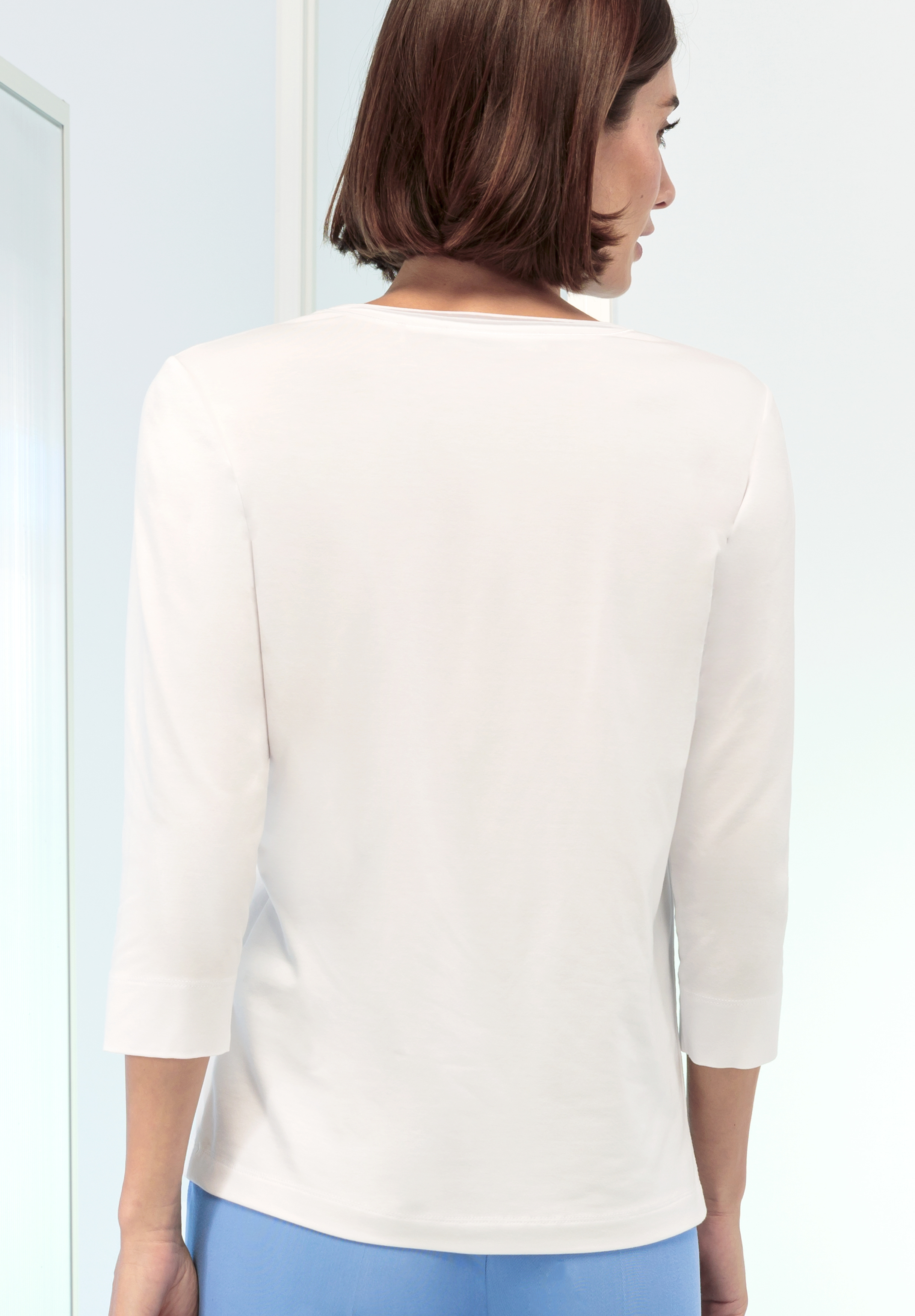 Von hinten ist eine Person mit kurzen braunen Haaren zu sehen, die ein weißes Hemd mit Dreiviertelärmeln und eine hellblaue Hose trägt und in einem Innenbereich vor einem hellen Hintergrund steht.