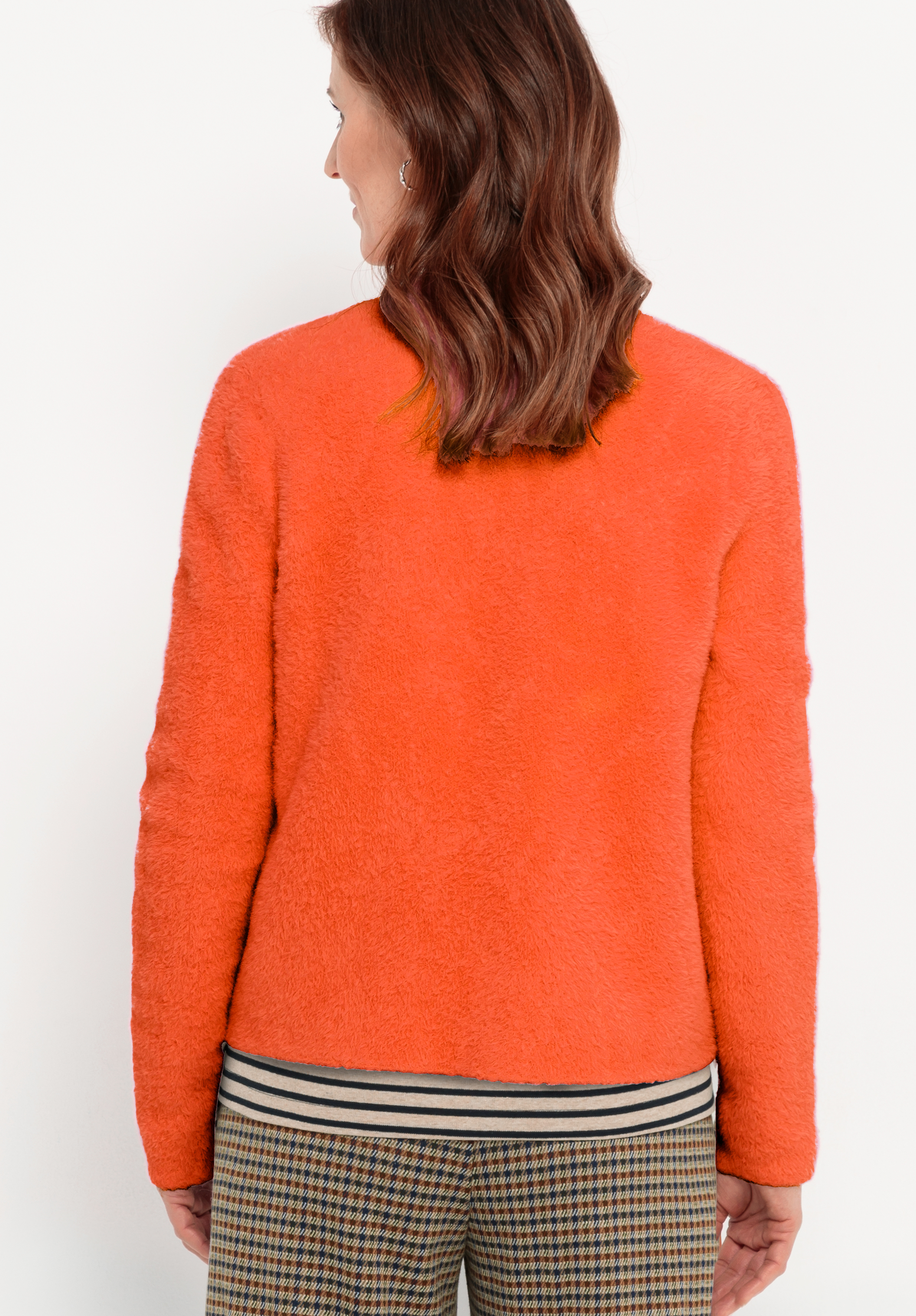 Eine Frau mit gewelltem braunem Haar, bekleidet mit einem orangefarbenen wuscheligen Pullover und einer karierten Hose mit gestreiftem Bund, steht mit dem Gesicht zur Kamera vor einem schlichten weißen Hintergrund.