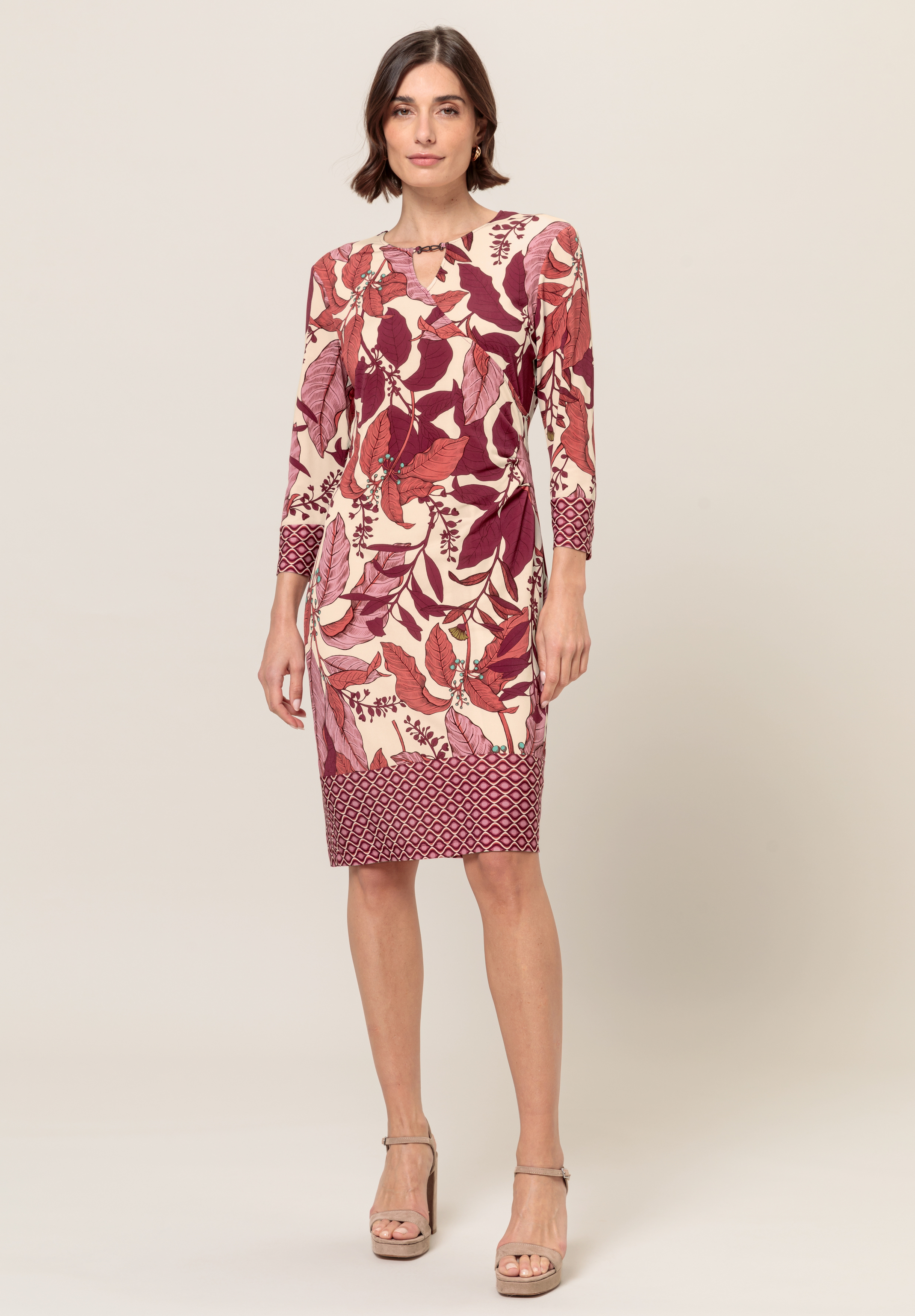 Eine Frau trägt ein knielanges, langärmeliges Kleid mit einem kräftigen rot-rosa Blattmuster und einer geometrischen Borte an Saum und Ärmeln. Sie hat kurzes braunes Haar und trägt beige Sandaletten mit hohen Absätzen.