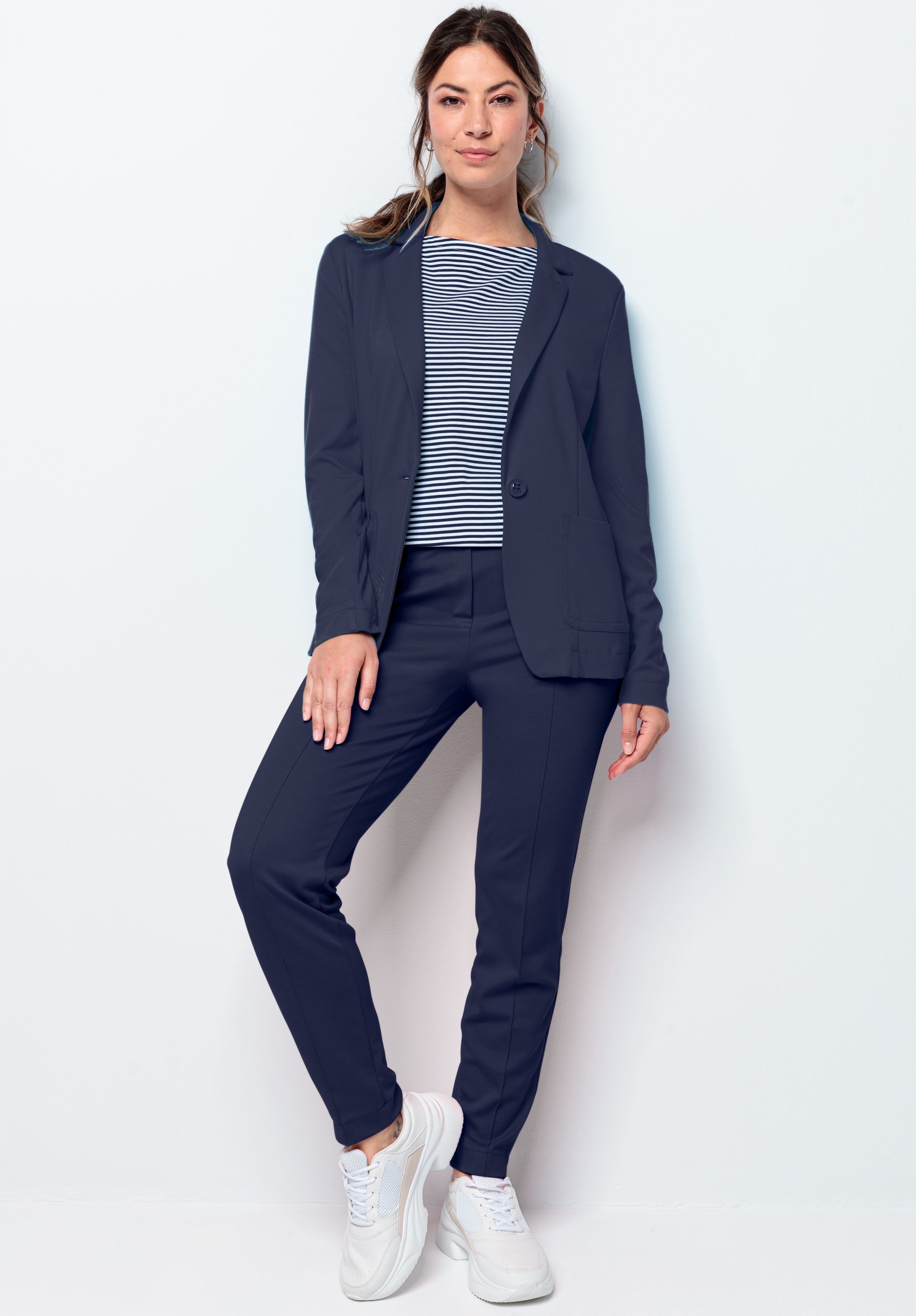Eine Frau steht vor einer weißen Wand. Sie trägt einen dunkelblauen Blazer und eine dunkelblaue Hose, ein gestreiftes dunkelblau-weißes Hemd und weiße Turnschuhe. Sie hat lange Haare zurückgekämmt, lächelt leicht und hat eine Hand in der Tasche.