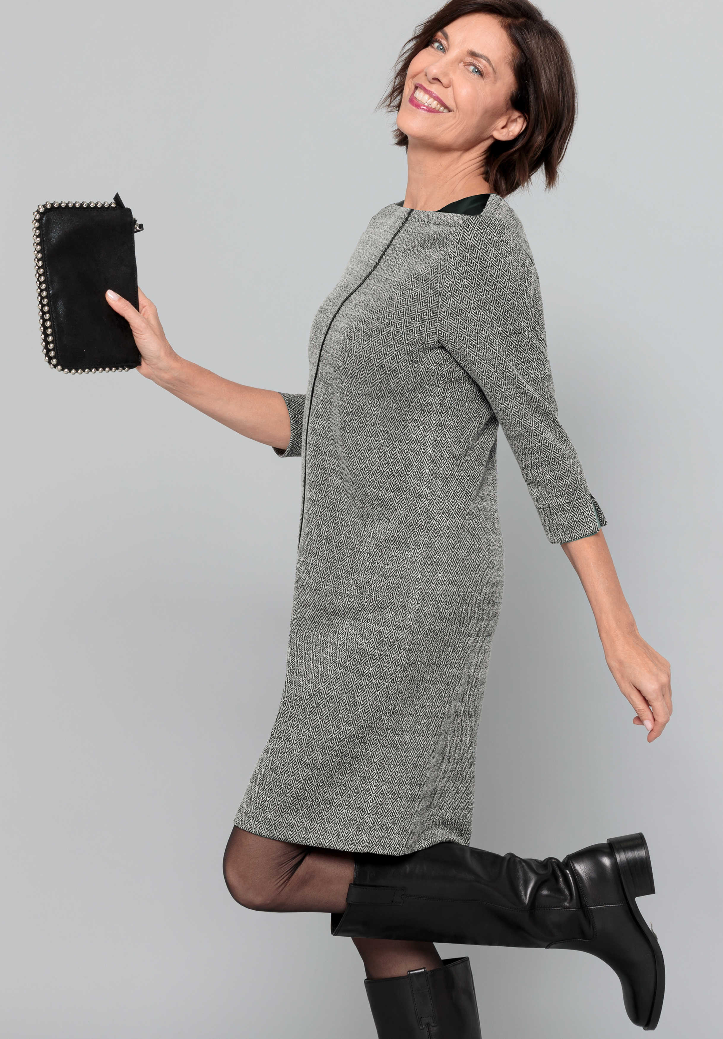 Eine Frau mit kurzen braunen Haaren, die ein graues, knielanges Kleid und schwarze Stiefel trägt und eine schwarze Clutch in der Hand hält, posiert verspielt auf einem Bein vor einem schlichten hellgrauen Hintergrund und lächelt in die Kamera.