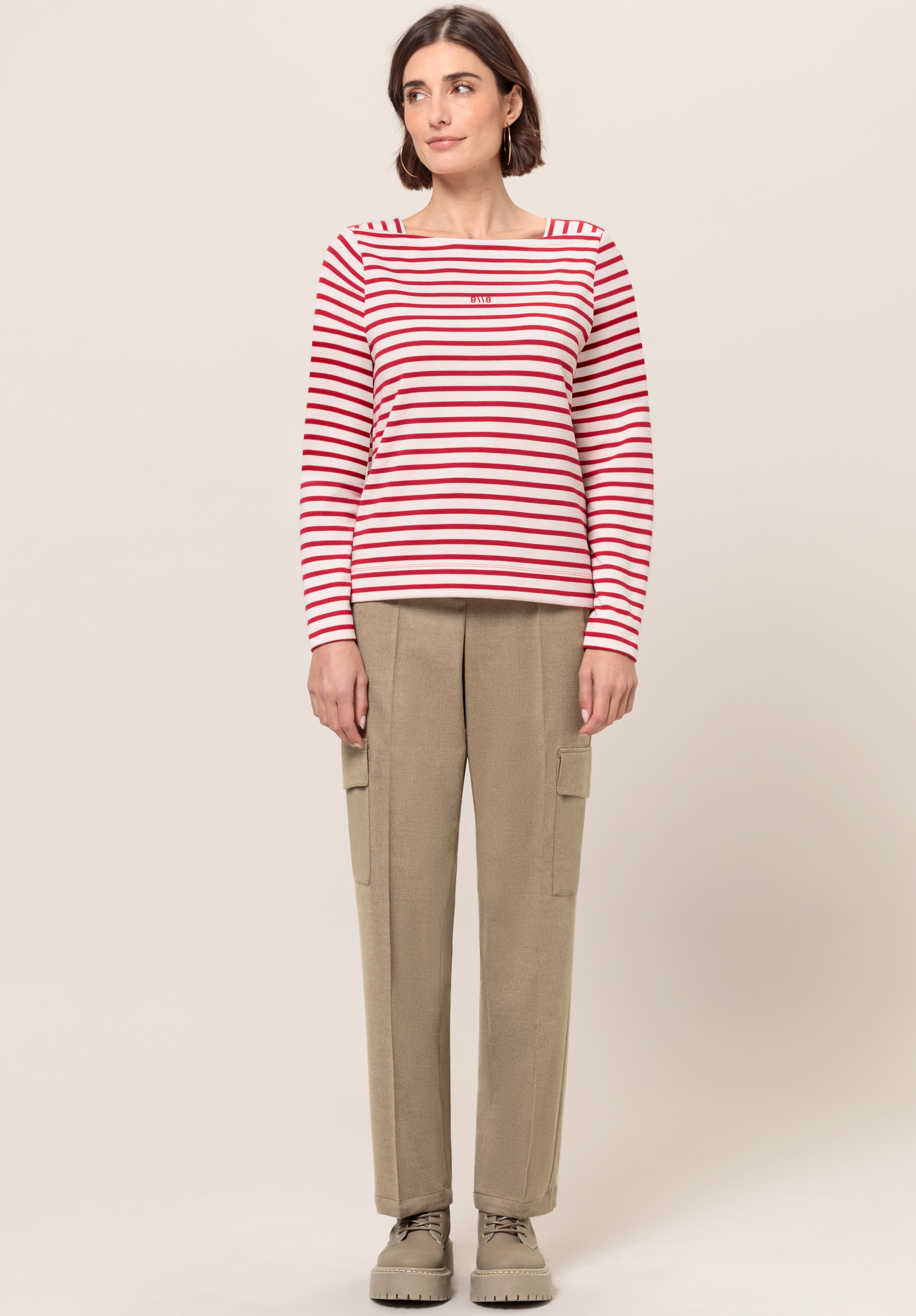Eine Frau steht vor einem schlichten Hintergrund. Sie trägt ein rot-weiß gestreiftes Langarmshirt, beige Cargohosen und beige Schnürschuhe. Sie hat kurze braune Haare und blickt mit neutralem Gesichtsausdruck leicht zur Seite.