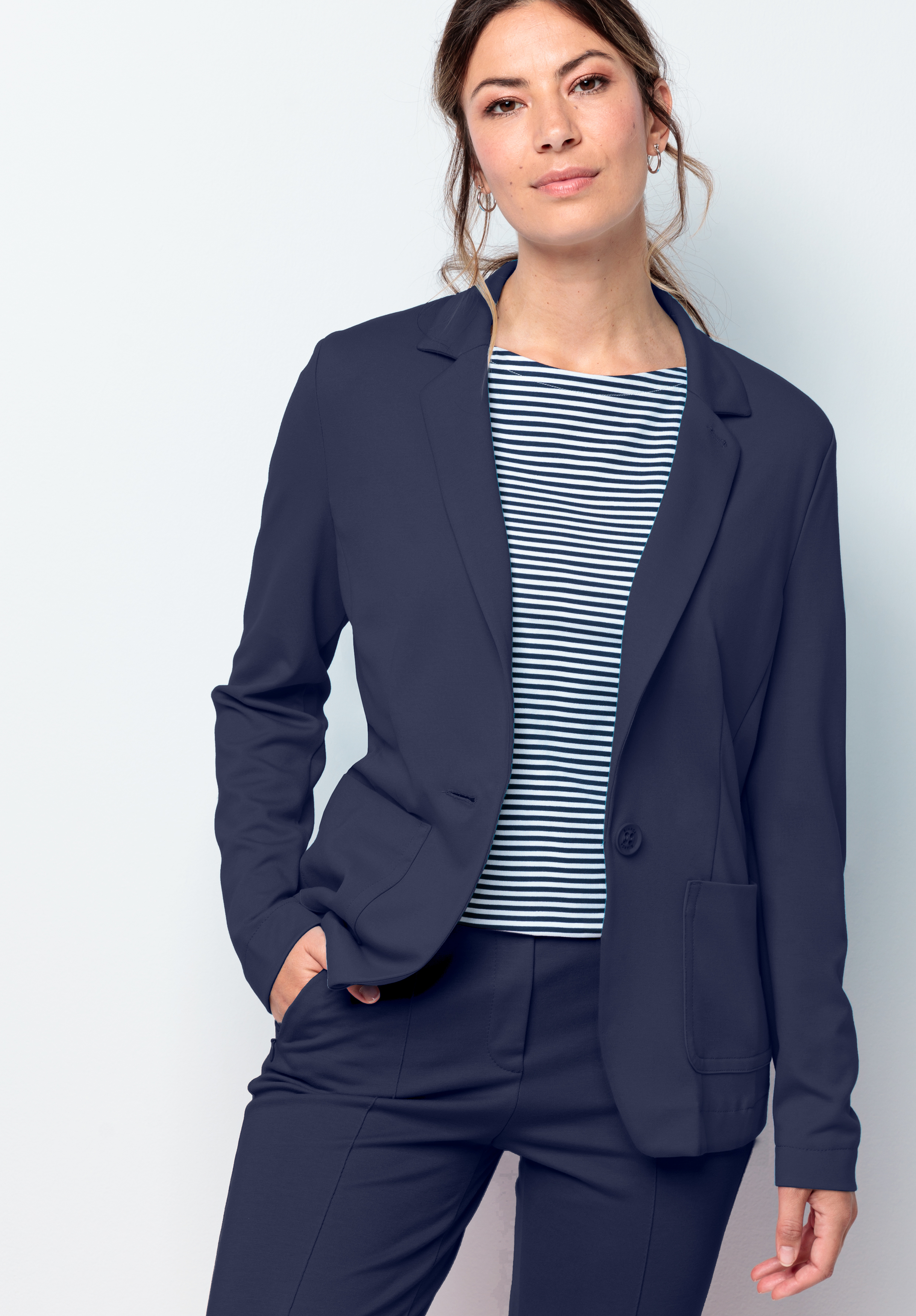 Eine Frau steht vor einem hellen Hintergrund. Sie trägt einen marineblauen Blazer und eine marineblau-weiß gestreifte Hose. Sie hat helle Haut, langes, zurückgekämmtes Haar und einen neutralen Gesichtsausdruck. Eine Hand steckt in der Tasche.