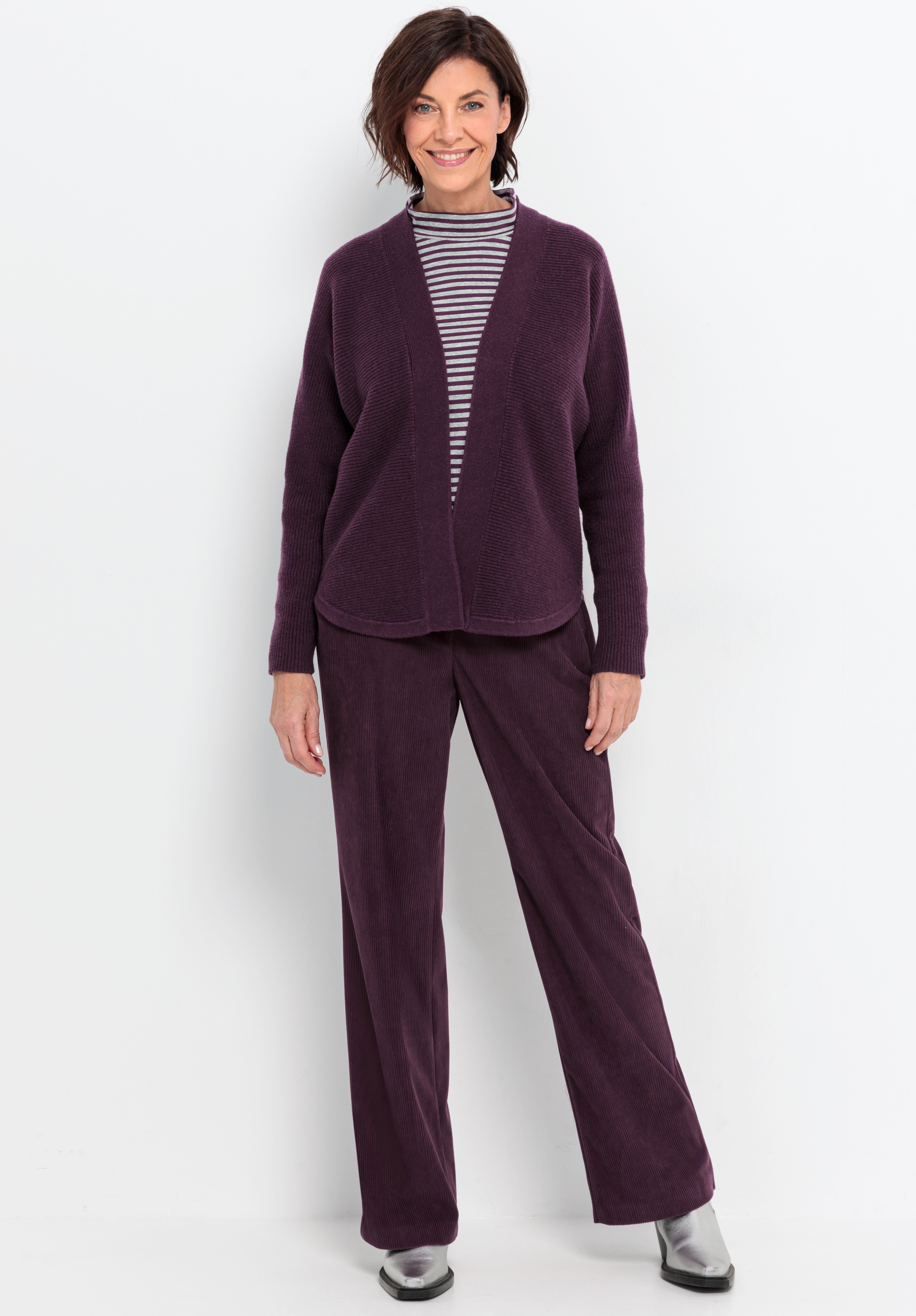 Eine Frau mit kurzen braunen Haaren steht lächelnd vor einem weißen Hintergrund und trägt eine lila Strickjacke, eine passende lila Hose, einen gestreiften Rollkragenpullover und silberne Schuhe.