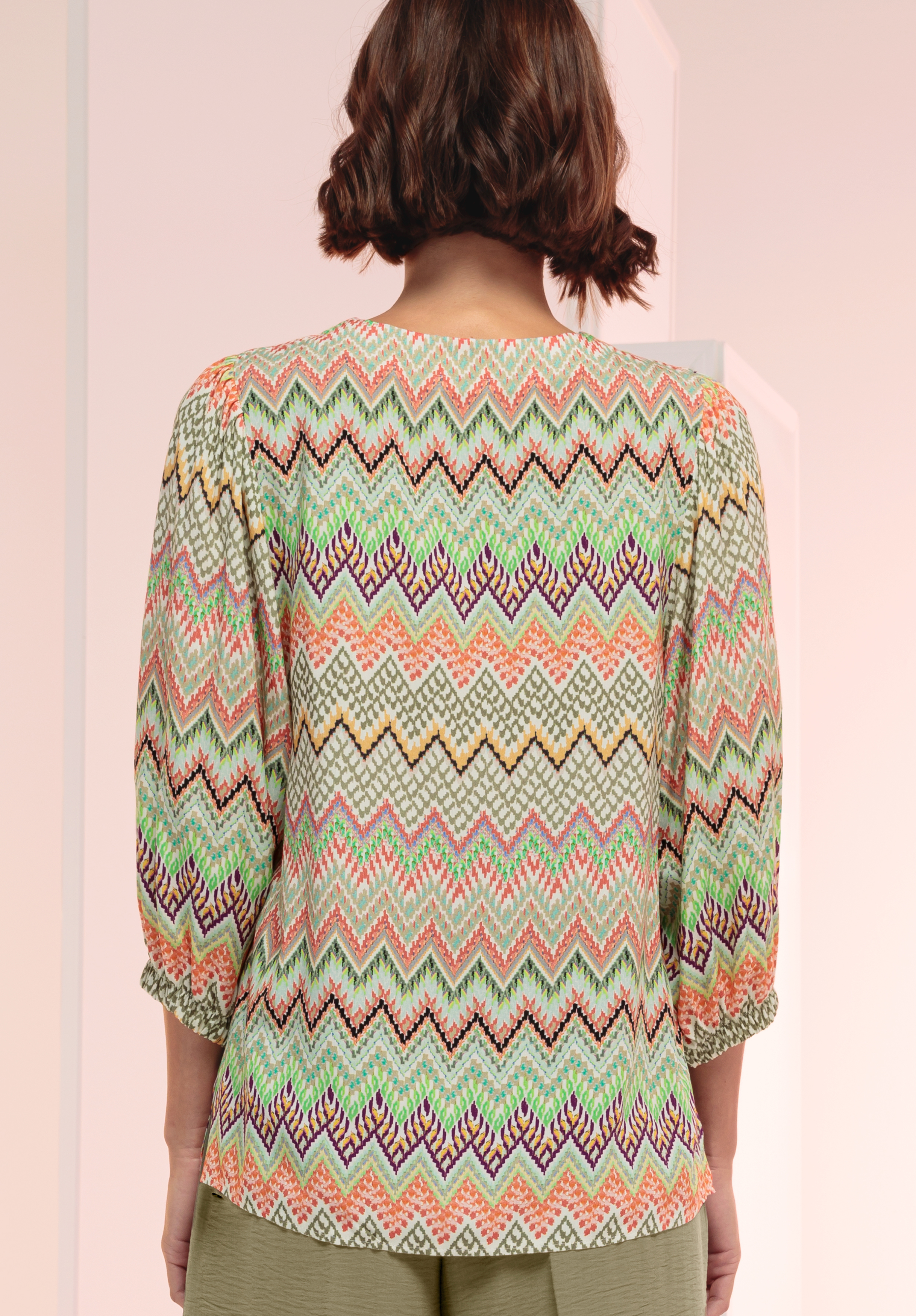 Eine Person mit kurzen braunen Haaren ist von hinten zu sehen. Sie trägt eine bunte Bluse mit Chevron-Muster und Dreiviertelärmeln sowie eine grüne Hose vor einem zartrosa-weißen Hintergrund.