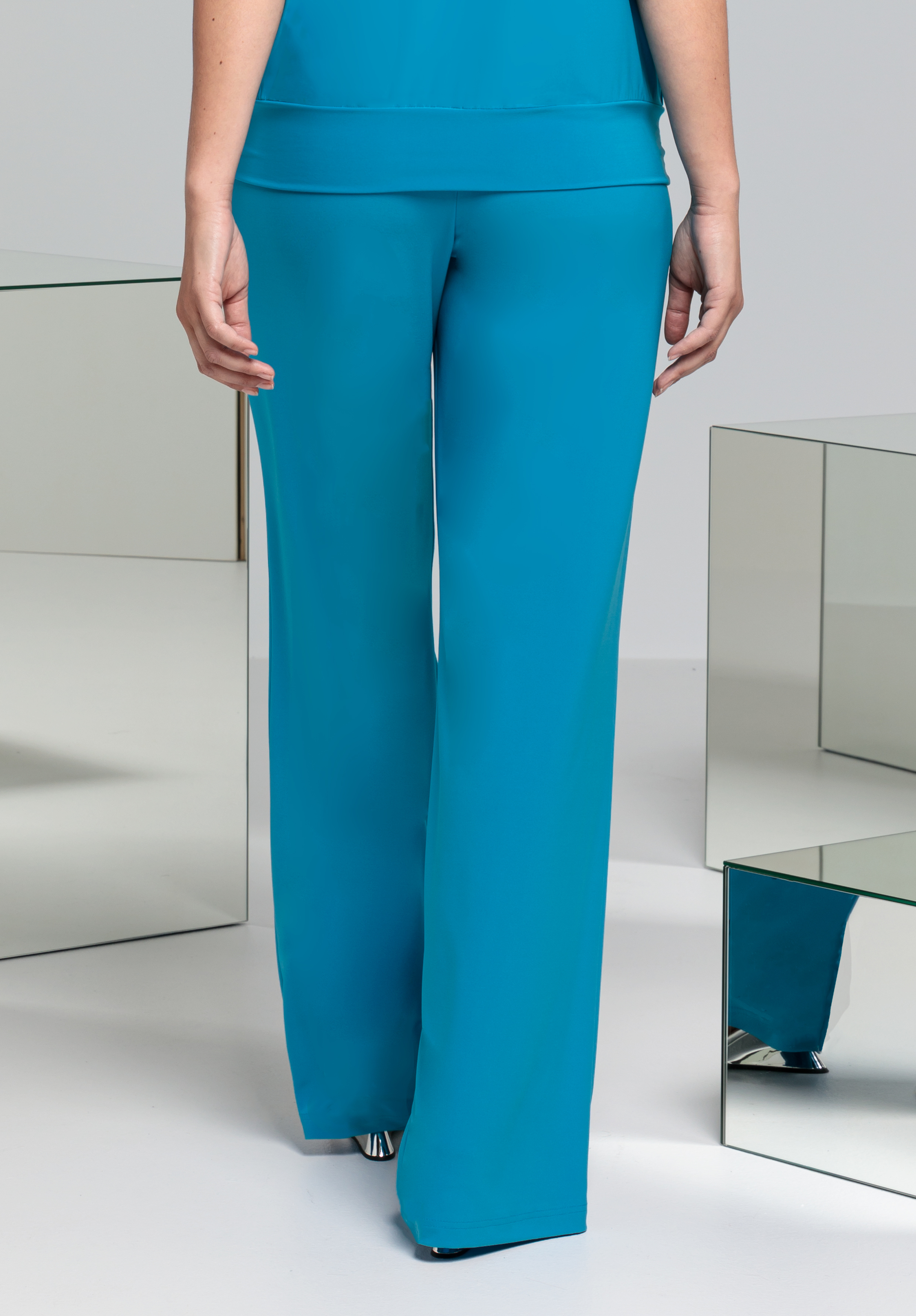 Eine Person in blaugrünen Hosen mit weitem Bein und einem dazu passenden Oberteil steht mit dem Rücken zur Kamera, umgeben von verspiegelten geometrischen Tischen auf einem hellen Boden.