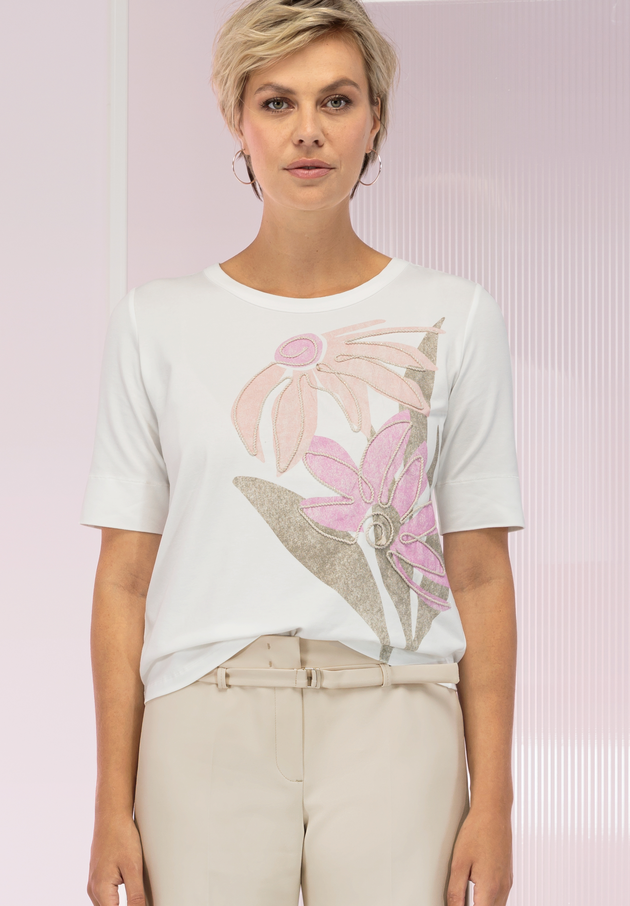 Eine Frau mit kurzen blonden Haaren trägt ein weißes T-Shirt mit rosa-beigem Blumenmuster, beige Hosen und Creolen und steht vor einem hellrosa Hintergrund.