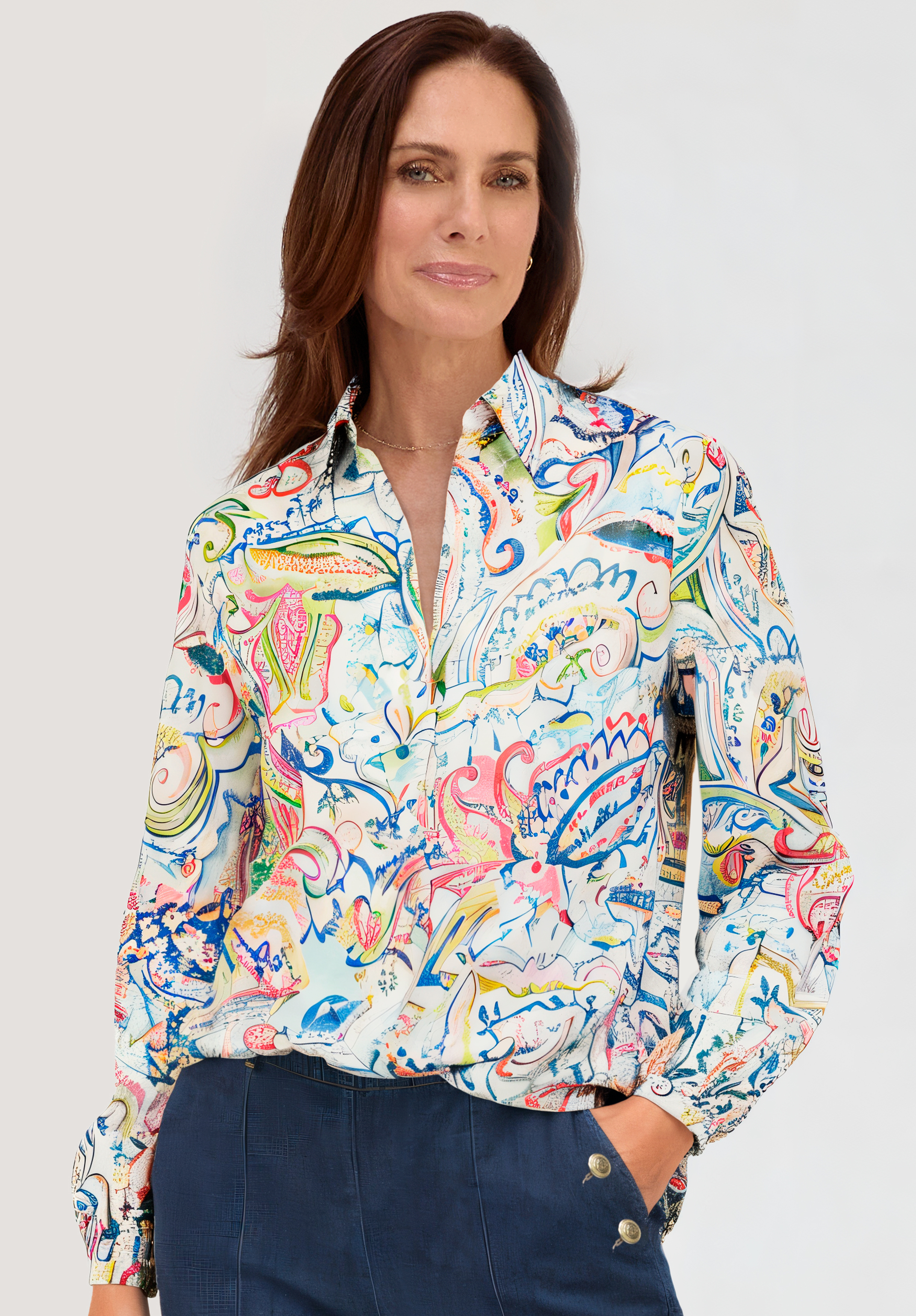Eine Frau mit braunen Haaren, die eine bunte, gemusterte Bluse mit Knöpfen und eine marineblaue Hose mit Knöpfen trägt, posiert selbstbewusst vor einem hellen Hintergrund.