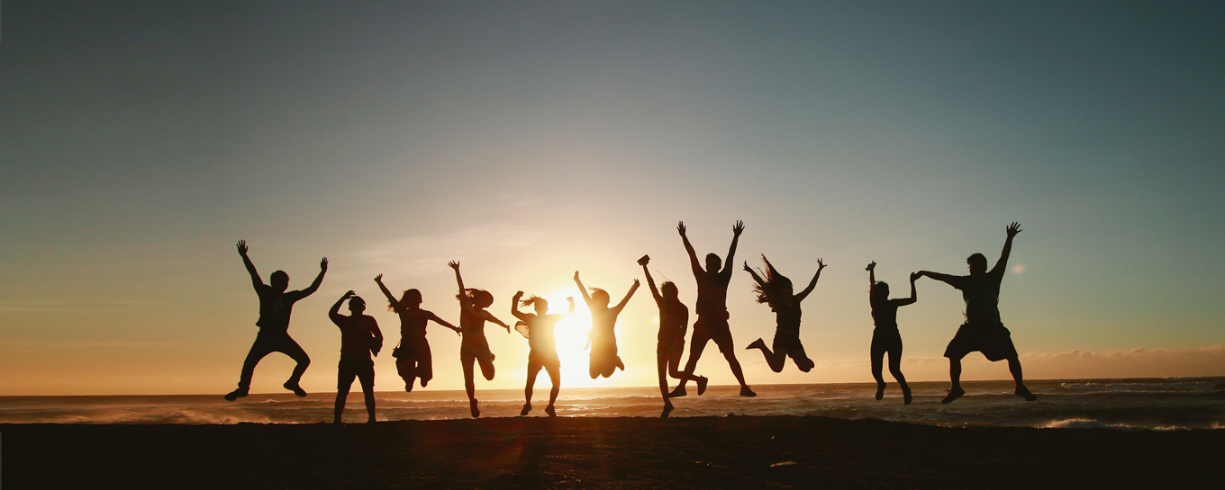 Eine Gruppe von Menschen springt bei Sonnenuntergang an einem Strand und streckt die Arme in die Luft. Die Silhouette hebt sich vor dem hellen Himmel und dem Meer ab.