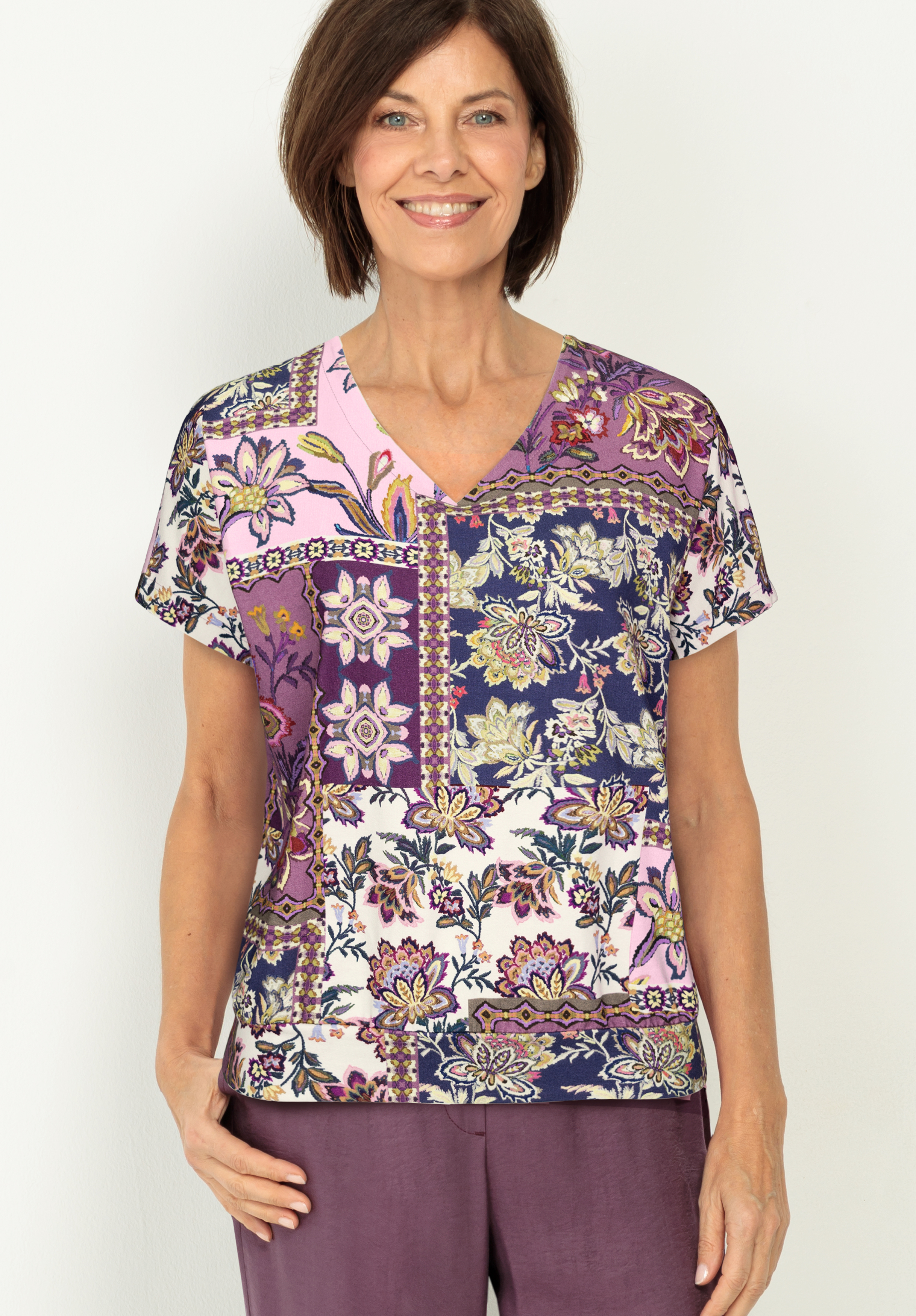 Eine Frau mit kurzen braunen Haaren lächelt, während sie eine bunte, blumengemusterte Bluse mit V-Ausschnitt in Lila-, Rosa- und Weißtönen und eine lila Hose vor einem hellen Hintergrund trägt.