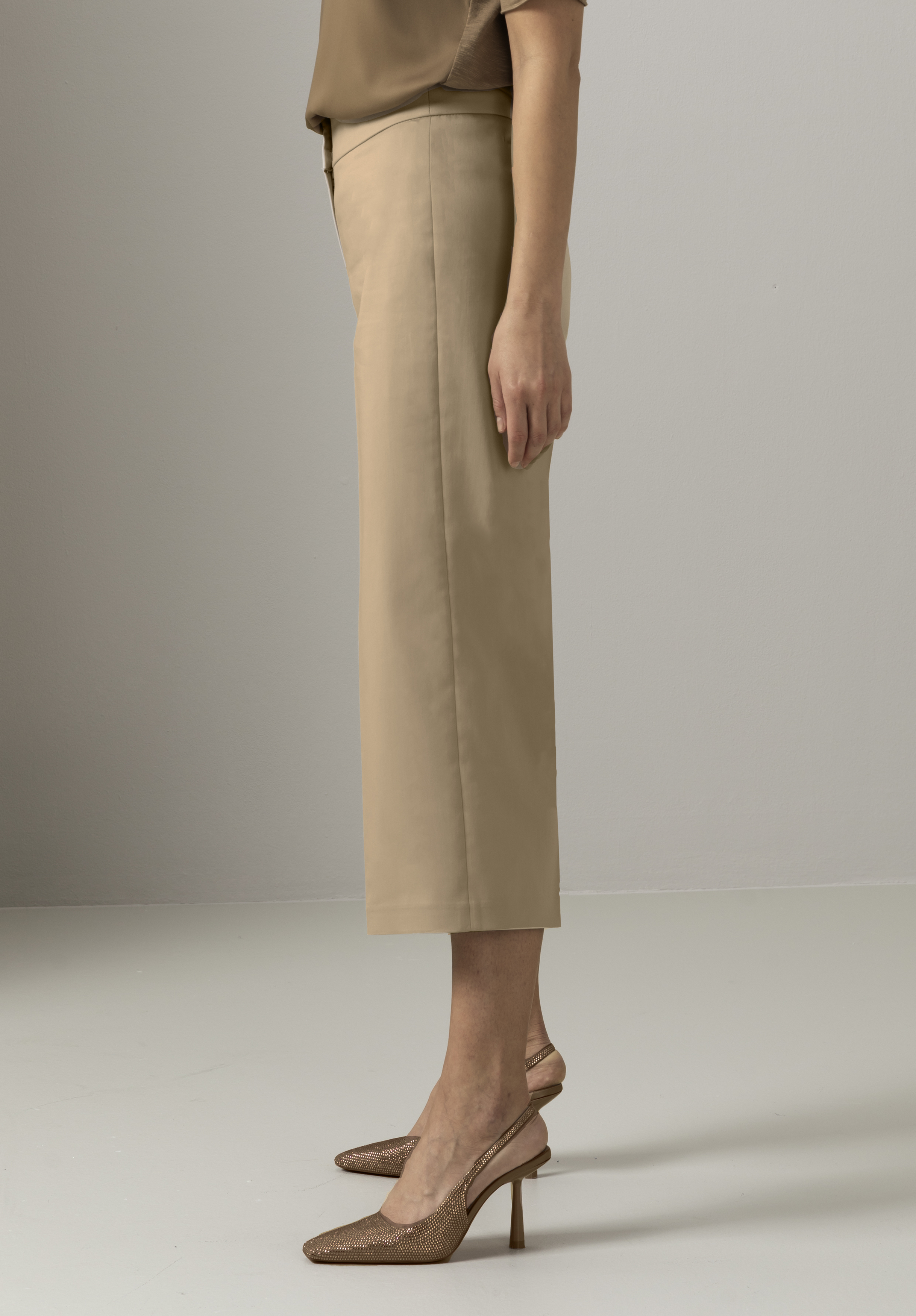 Eine Person steht im Profil und trägt kurze beige Hosen, eine braune Bluse und braune Slingback-Schuhe mit hohen Absätzen in einer minimalistischen Umgebung in neutralen Tönen.