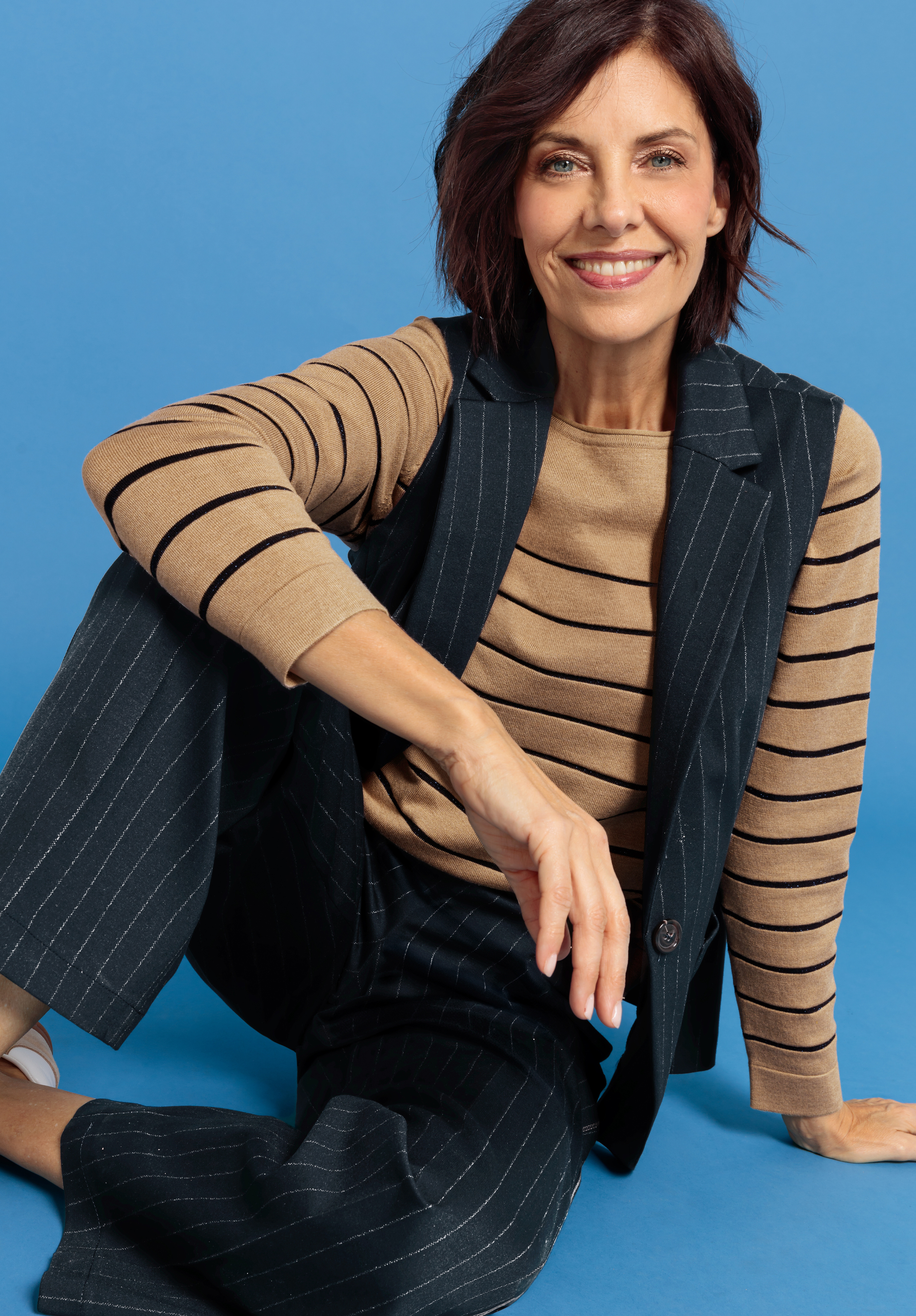 Anzugweste ANDRA Eine Frau mit kurzen braunen Haaren lächelt, während sie vor einem blauen Hintergrund sitzt. Sie trägt einen hellbraunen Pullover mit schwarzen Streifen und einen dunklen Nadelstreifenanzug mit Weste und Hose.